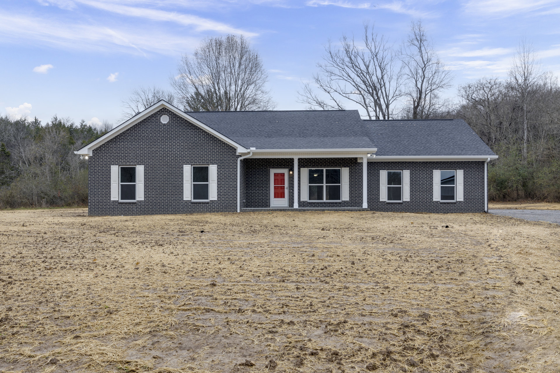 Brick house with red door and white shuttered windows, surrounded by a large dirt yard and mature trees under a cloudy sky