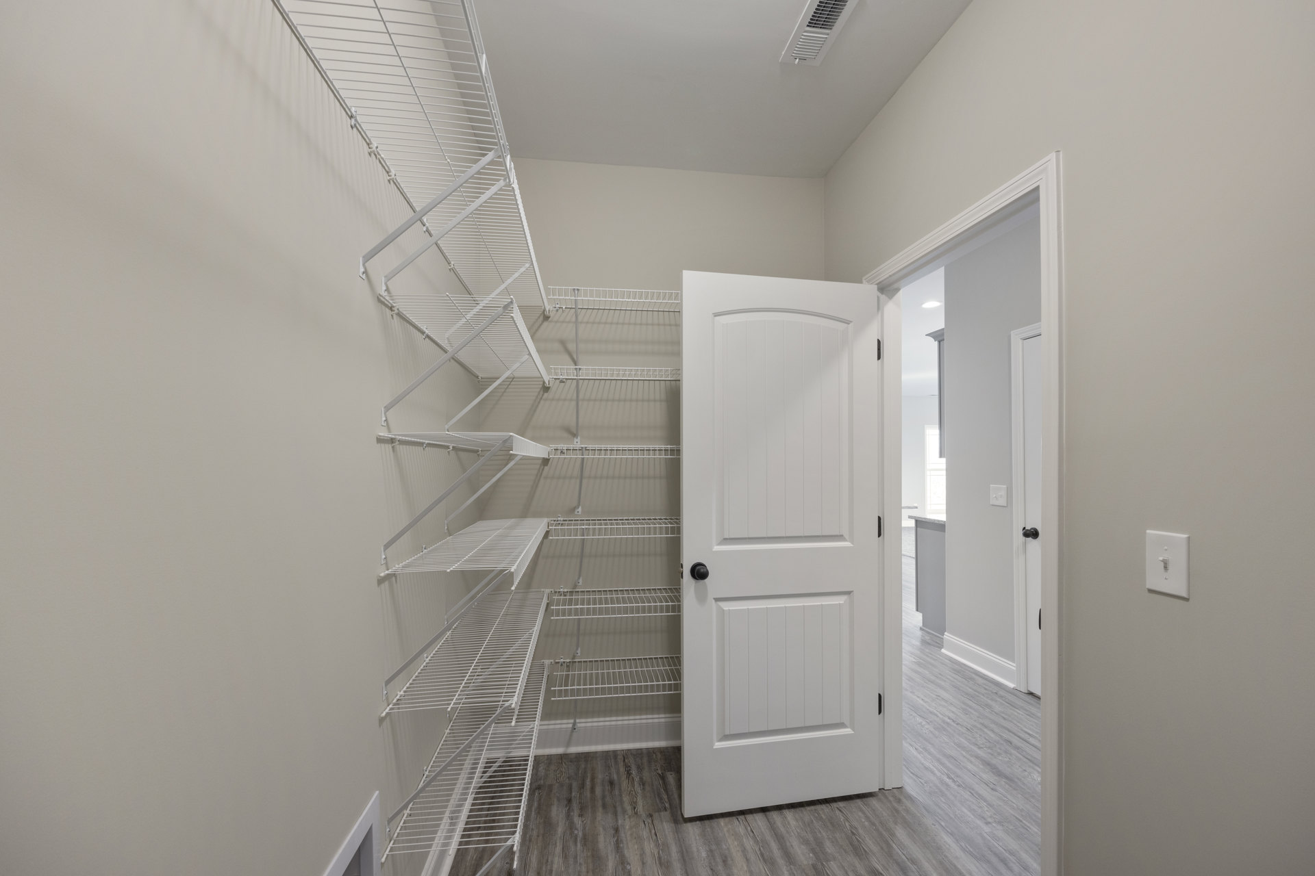 White paneled door with black knob, open to reveal built-in white shelves inside a closet; light switch on adjacent plaster wall, hardwood floor, white ceiling.