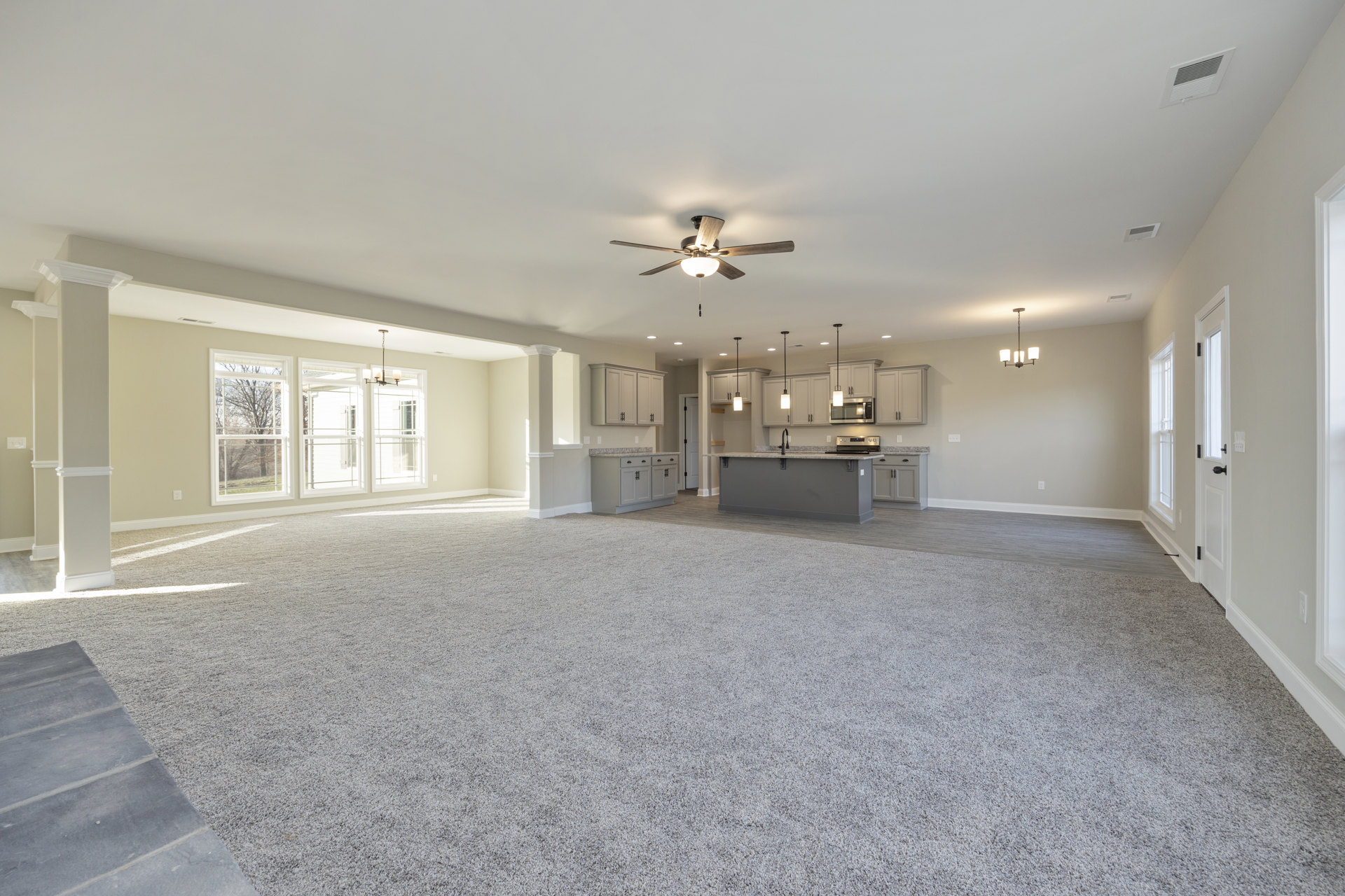 Spacious open-plan room featuring light-colored flooring, a modern kitchen with white cabinetry and a central counter, ceiling fan with integrated light, large window overlooking