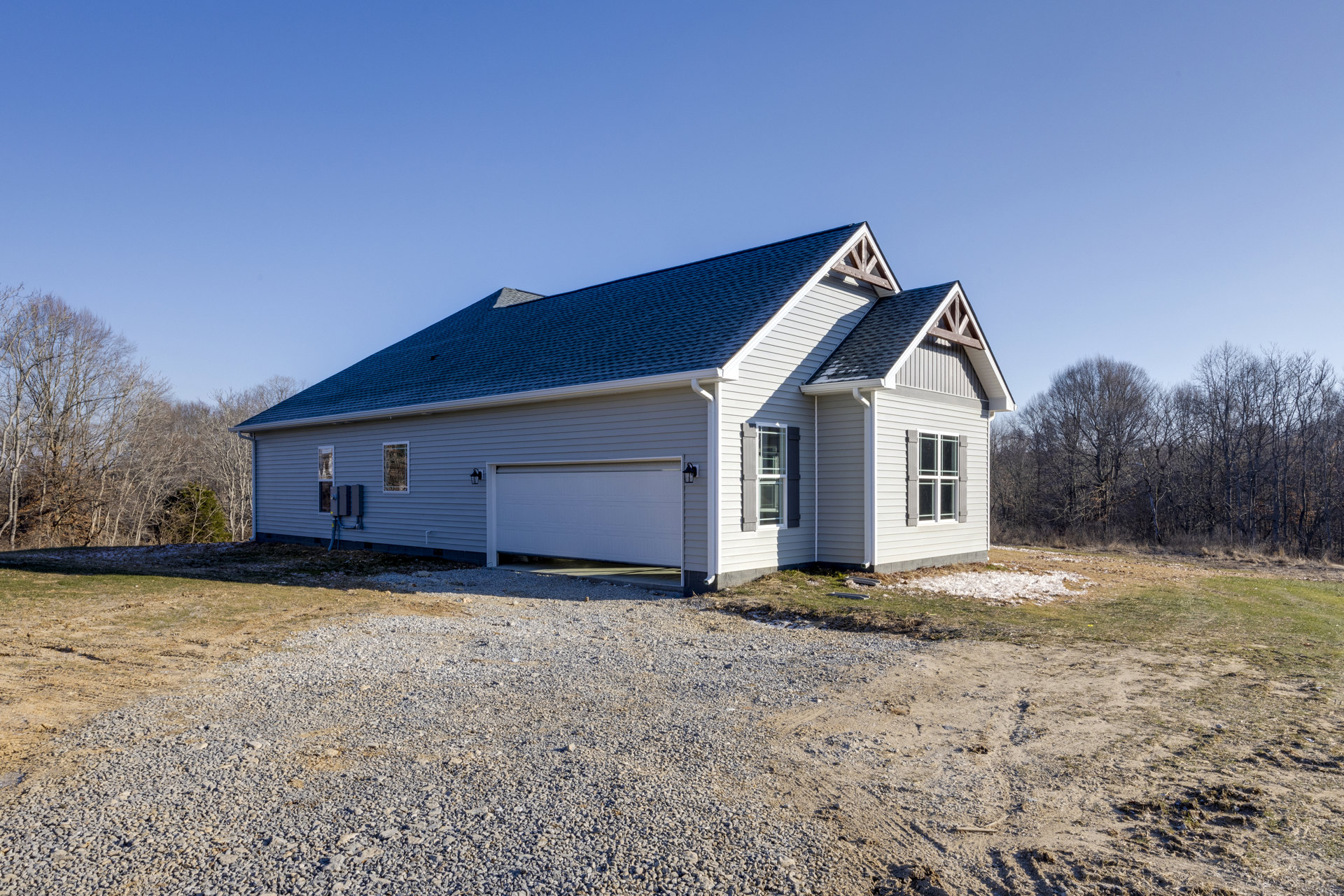 Two-story house with blue gabled roof, attached garage with white door, gravel driveway, large windows, surrounded by trees