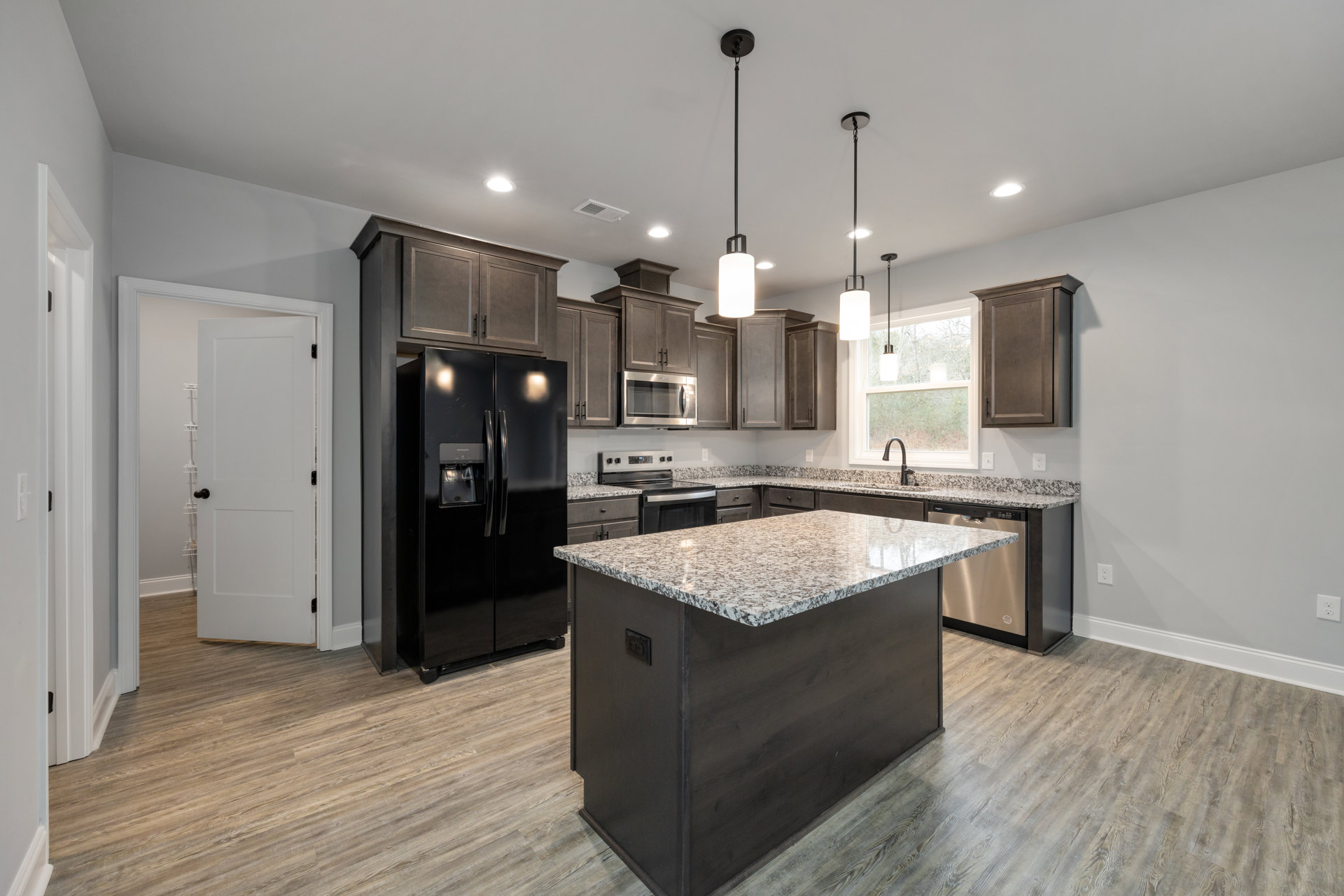 Spacious kitchen featuring a large granite island, black double-door refrigerator, brown cabinets with black handles, built-in microwave, white door with black handle, tile