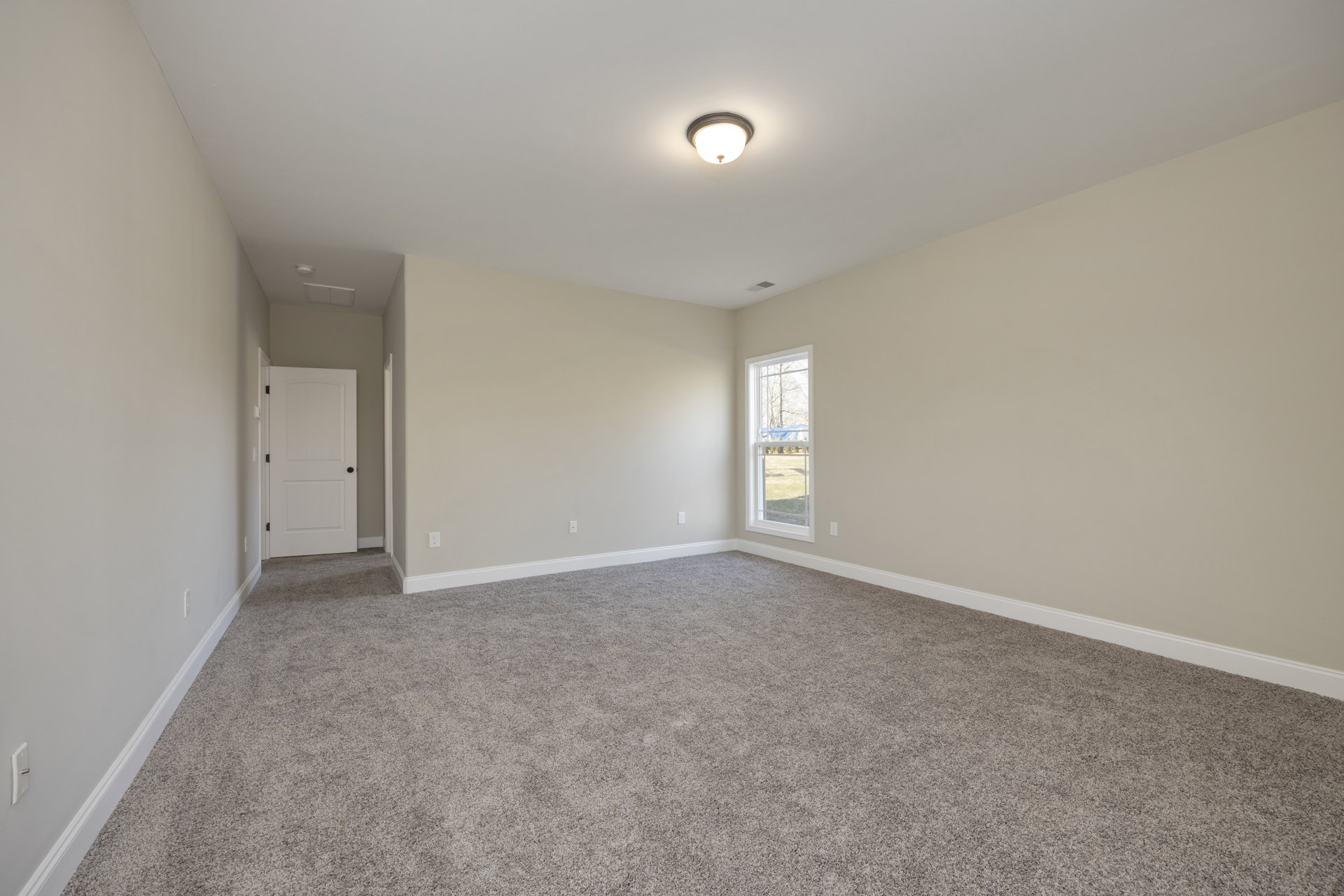 Carpeted room with ceiling light fixture, white door with black knobs, large window overlooking yard and trees, white walls, crown molding