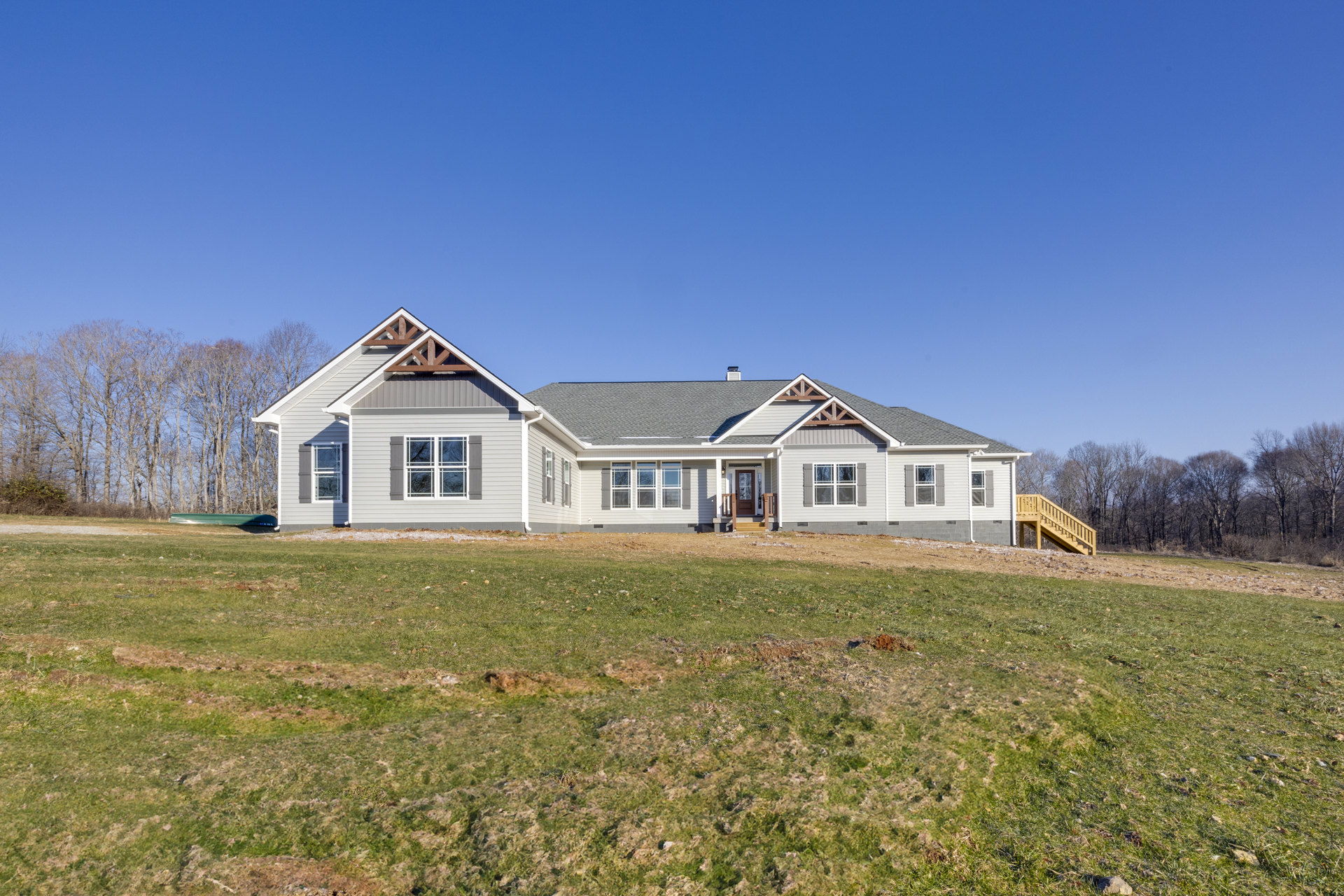 Two-story farmhouse with wide covered porch, wooden exterior stairs, large green lawn, mature trees in background, white-framed windows, gray roof under clear blue sky