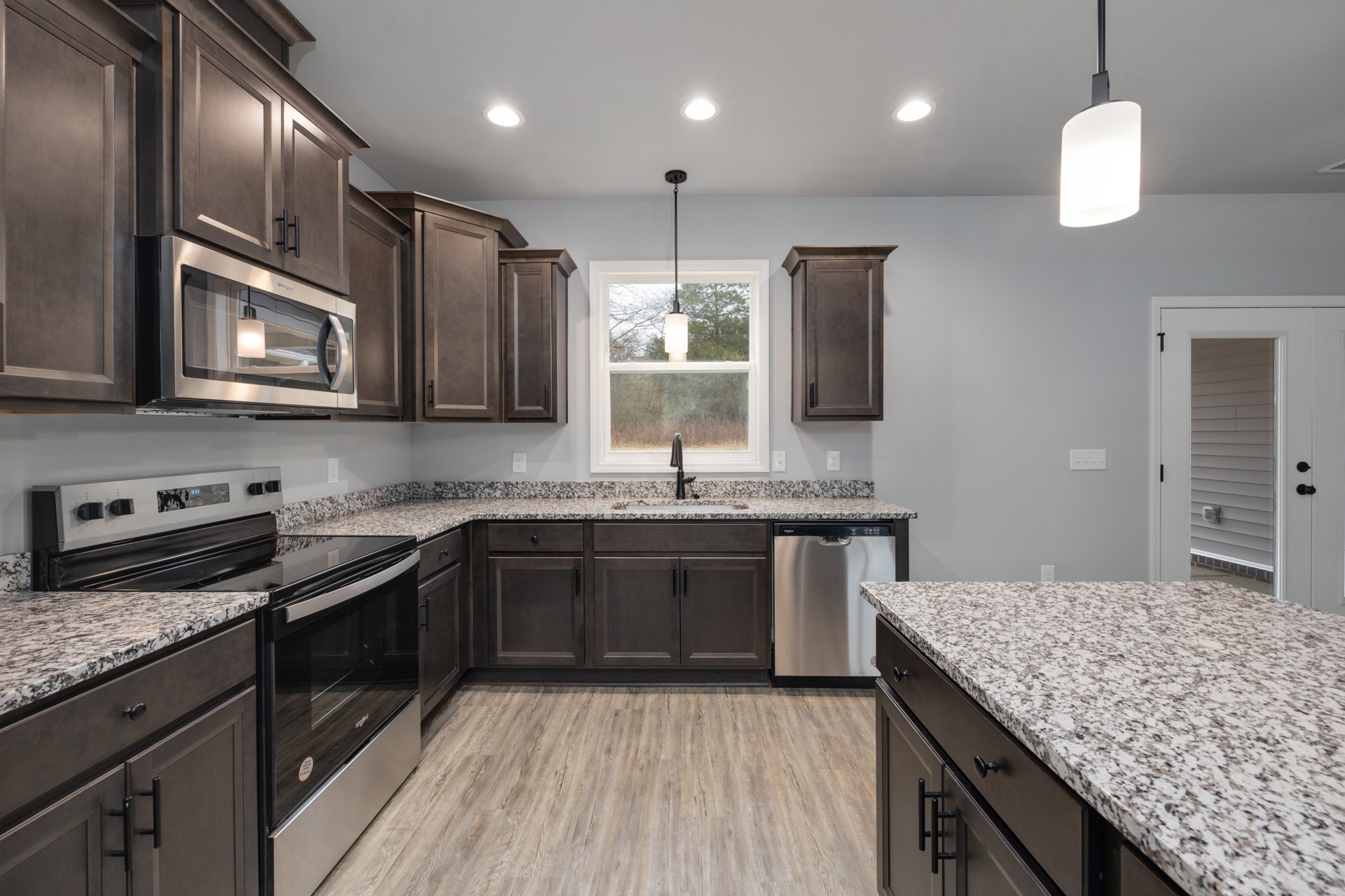 Granite kitchen countertops with black and white speckled surface, wooden cabinetry, stainless steel microwave, sink beneath a window, recessed lighting above