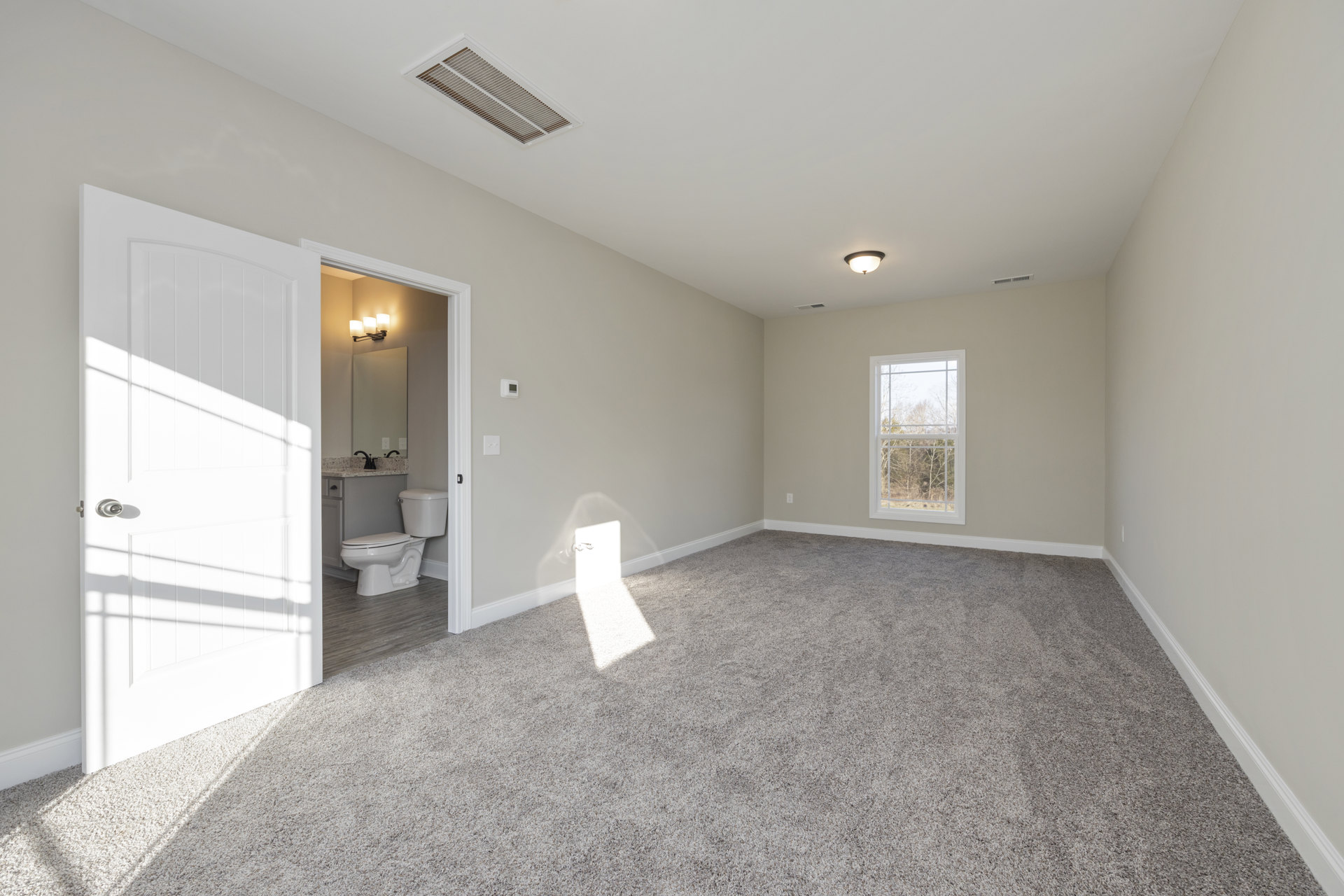 Bathroom with two white toilets, tile flooring, white walls, window showing trees outside, white door with silver knob, natural light streaming through wall