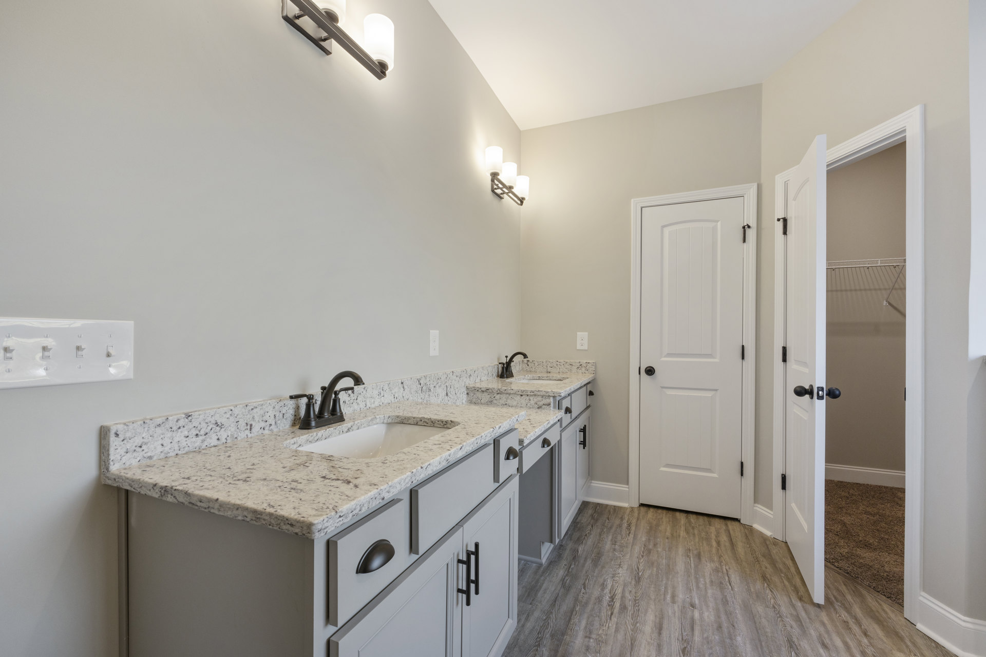 Bathroom with marble countertops, dual sinks with black faucets, wood flooring, white cabinetry, black door knob, and a white light switch on the wall