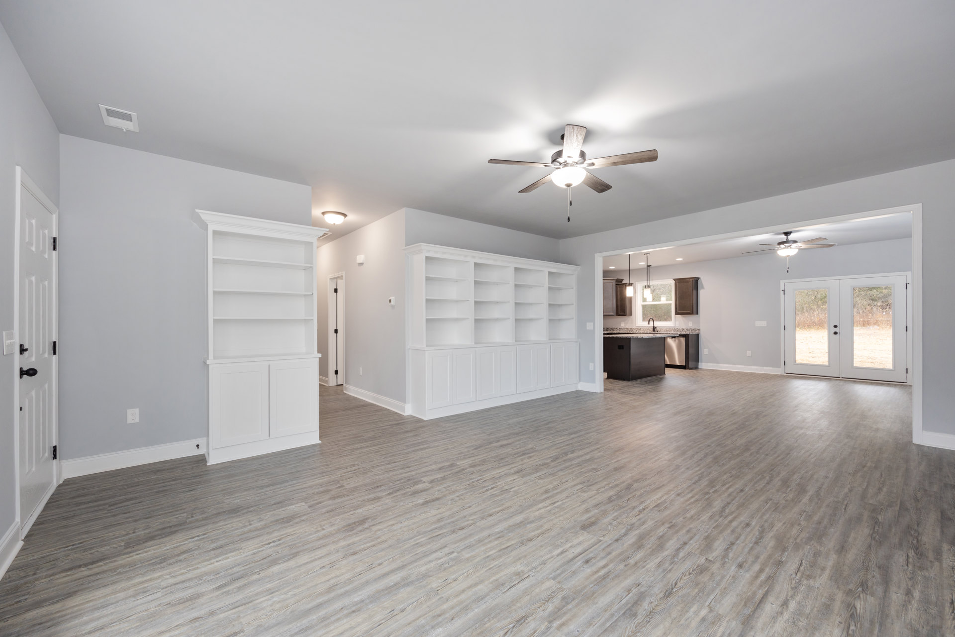 White built-in shelves along one wall, wood flooring, double glass-paneled doors, plaster walls, ceiling fan with light fixture.