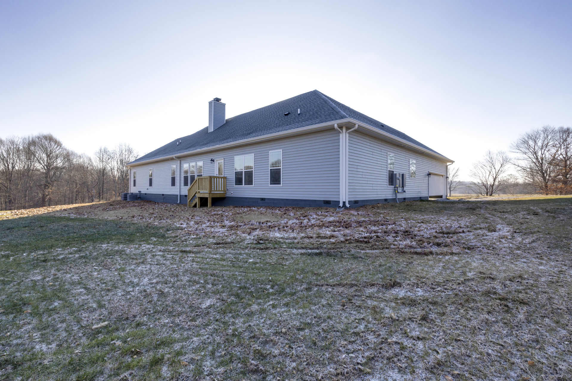 Two-story house with white-framed windows, wooden deck, and chimney, surrounded by a snowy grass field and bare trees under a winter sky