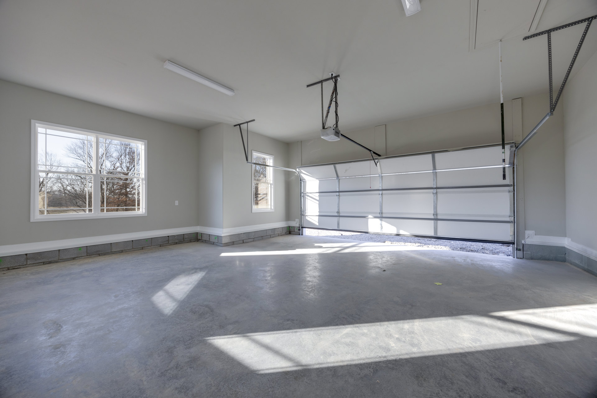 Spacious room with smooth white plaster walls, large windows overlooking trees, concrete flooring, and a garage door; white ceiling light fixture visible