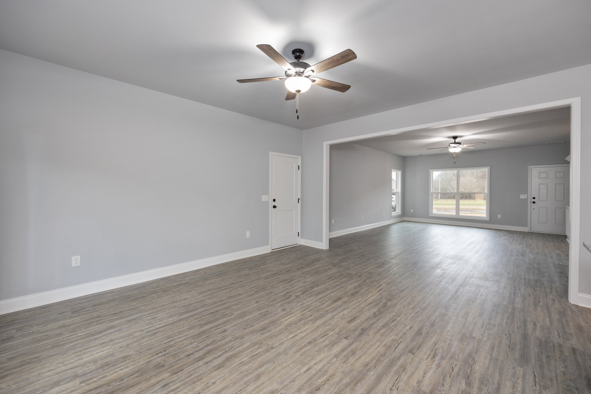 Ceiling fan with light fixture, wood flooring, white door with black handles, window overlooking yard and trees, white plaster walls