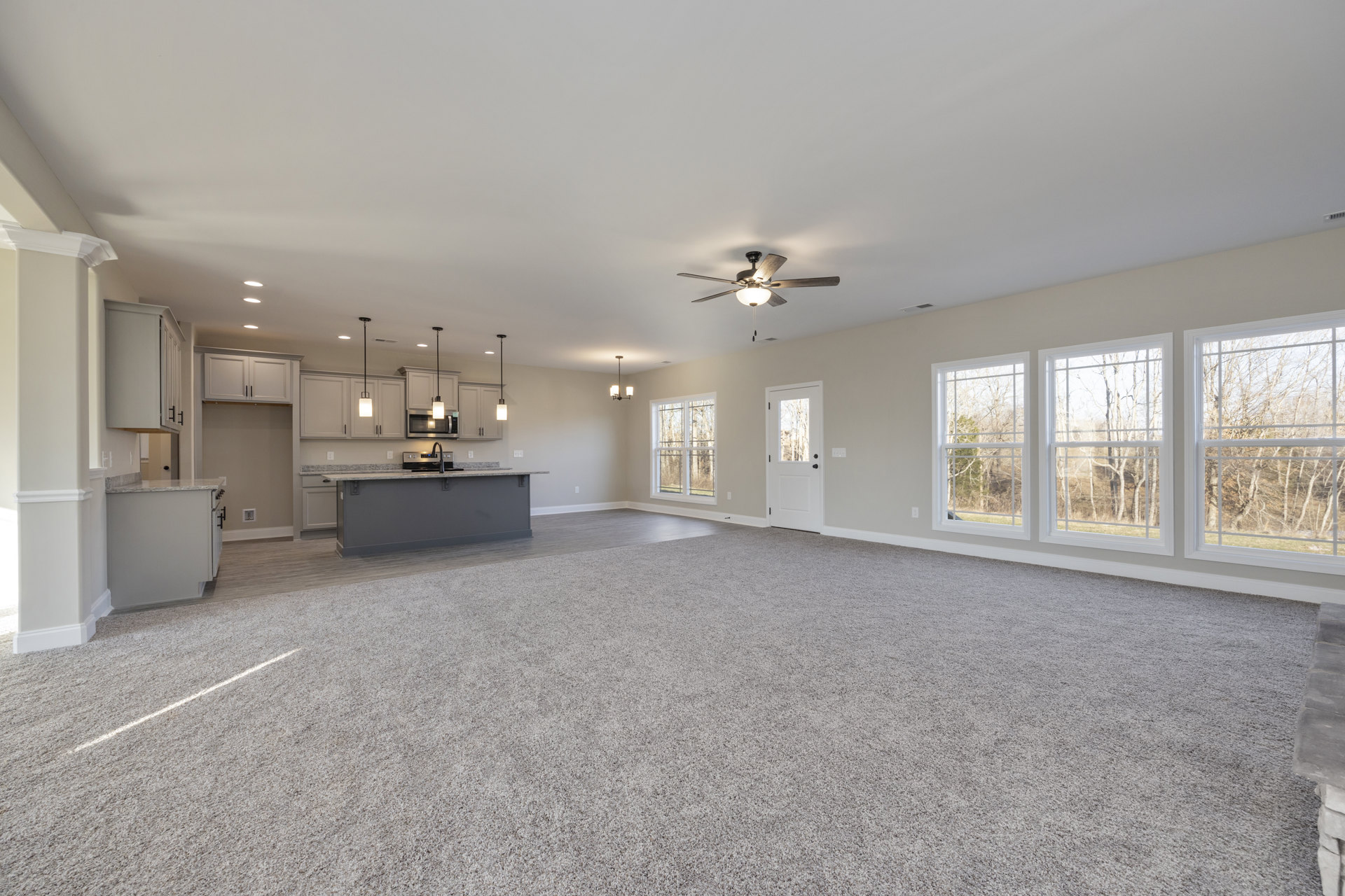 Open-concept room featuring a kitchen with tile flooring, multiple windows with views of trees, a white door with glass panel, and a ceiling fan with light fixture