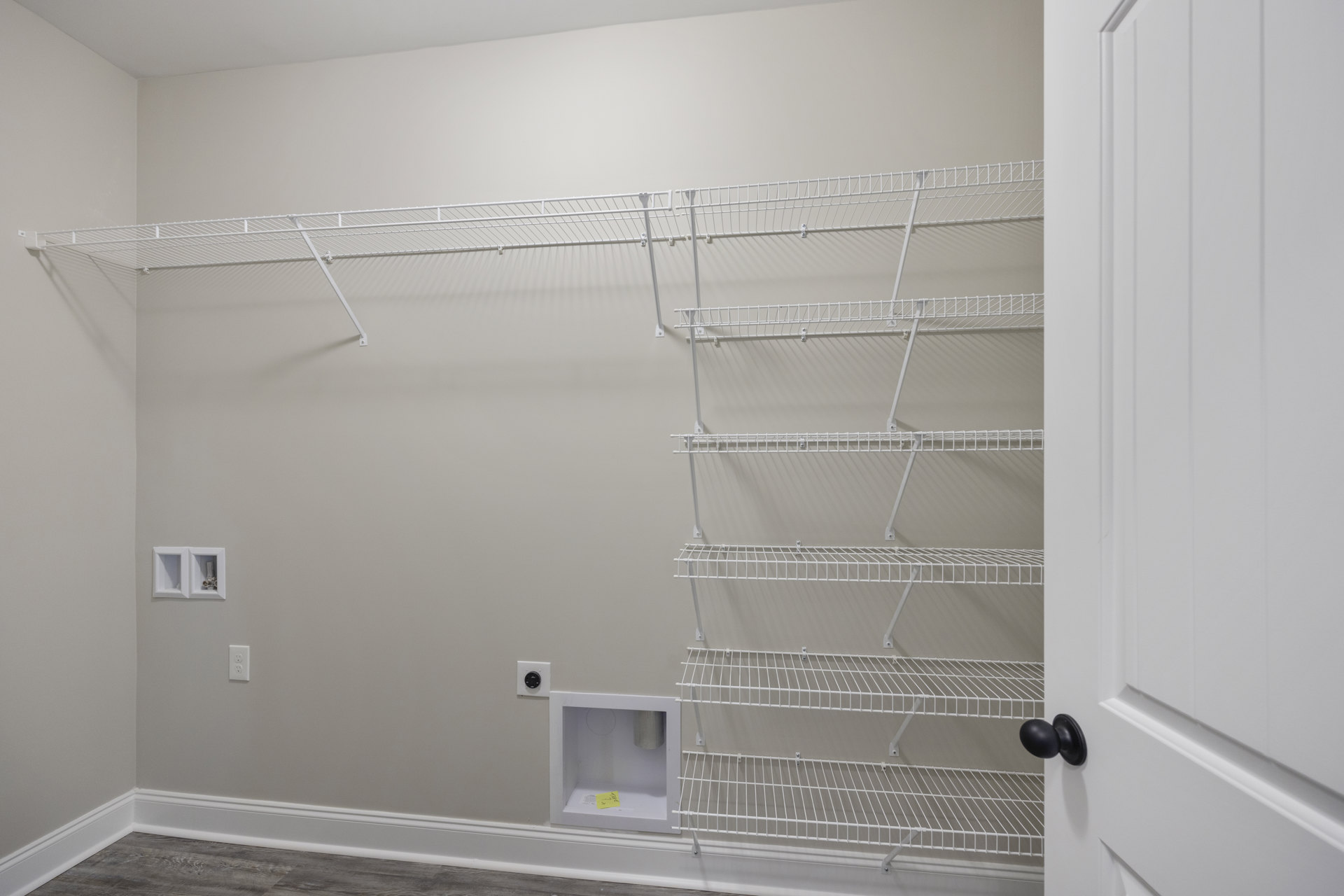 White wire shelving mounted on a plaster wall, white box with yellow paper on top, white electrical outlet with exposed wires, black door knob on a closed white door.