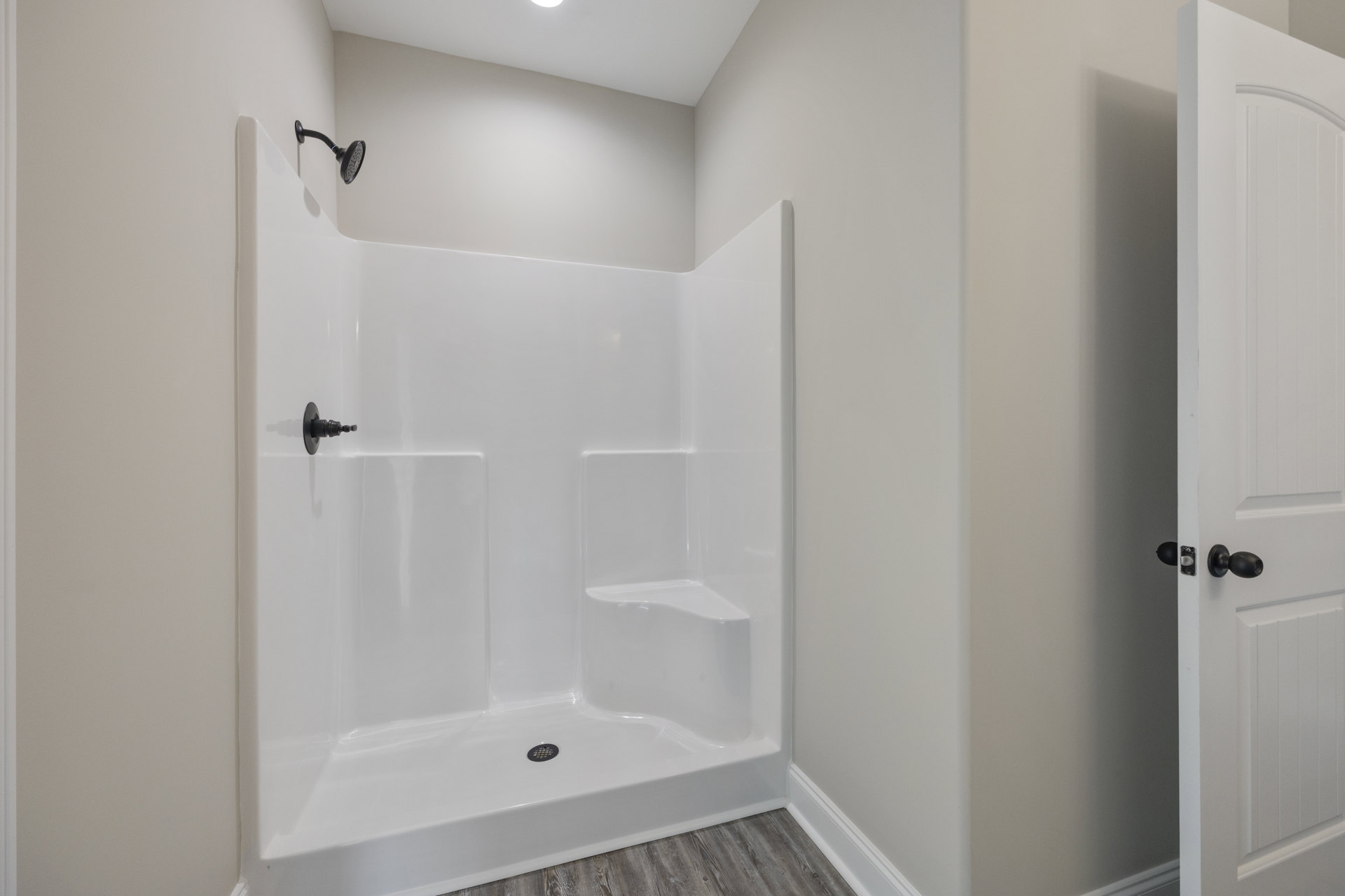 Black shower head with matching arm mounted on white tiled wall, white paneled door adjacent, shower floor features central drain, minimalist bathroom design with plaster ceiling