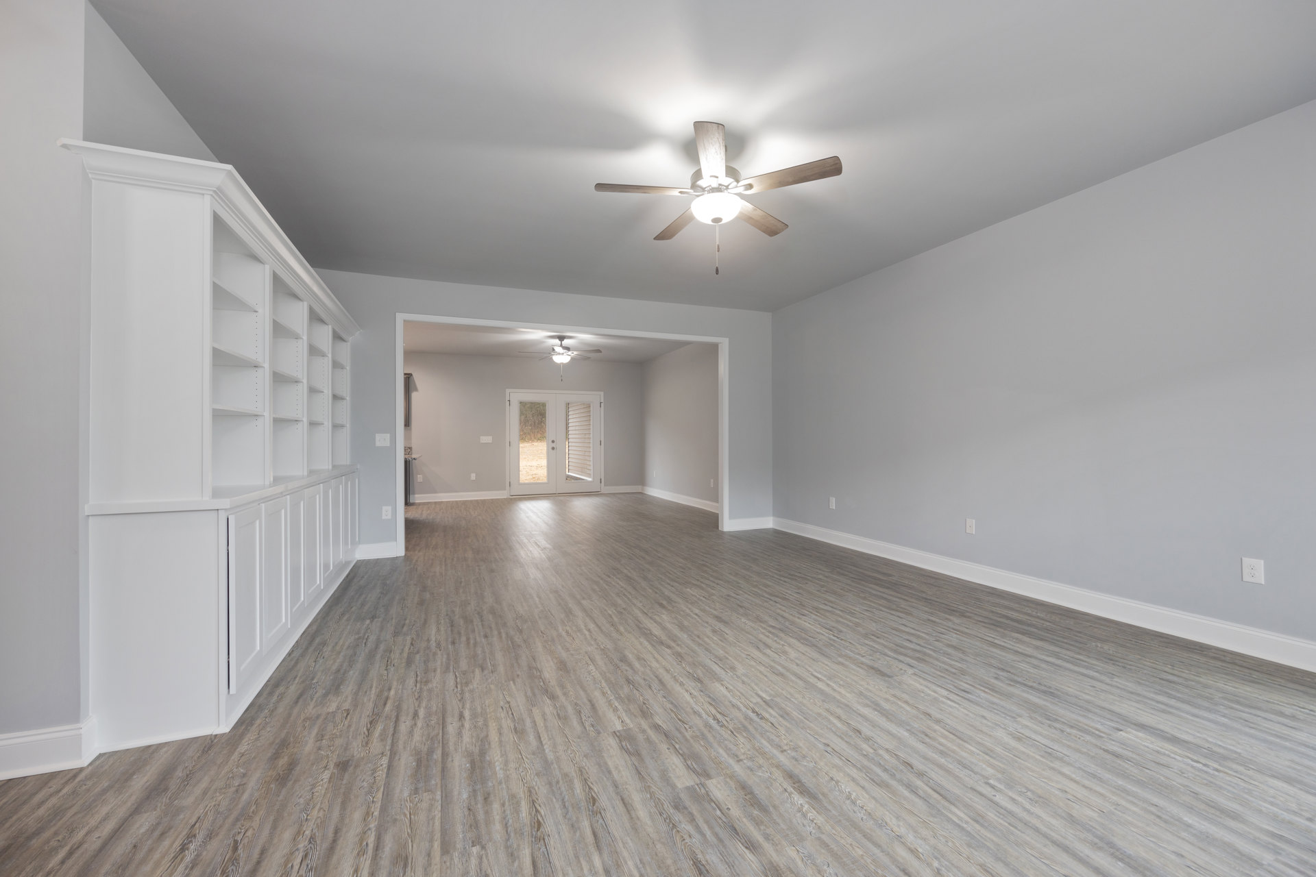 Ceiling fan with light fixture above wood laminate flooring, built-in white shelves along wall, double glass-paneled doors, smooth plaster ceiling and walls
