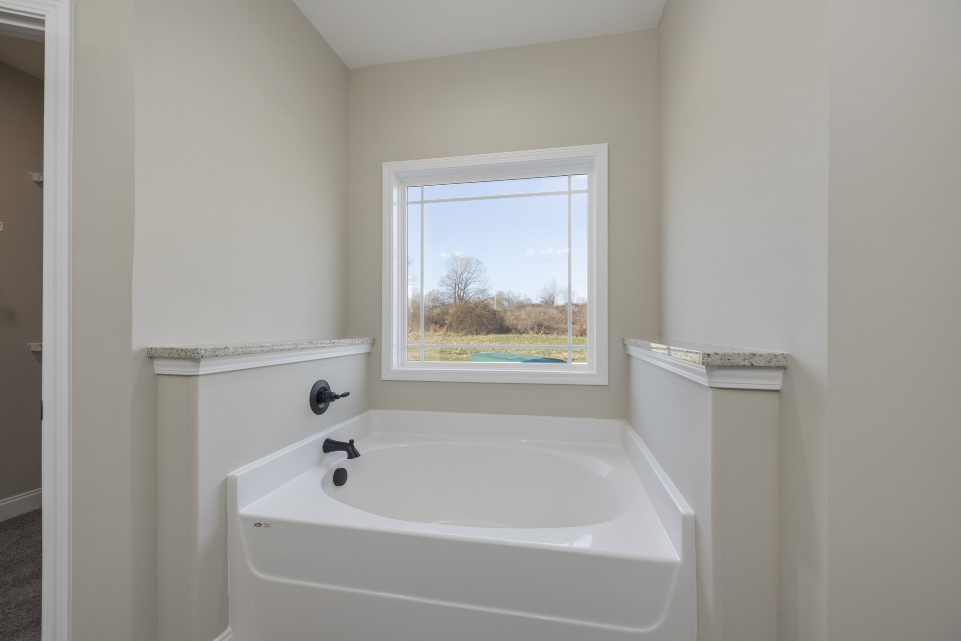 Freestanding white bathtub with black faucet beneath a large window overlooking a field and trees, surrounded by light tile walls and a close-up of a wall switch
