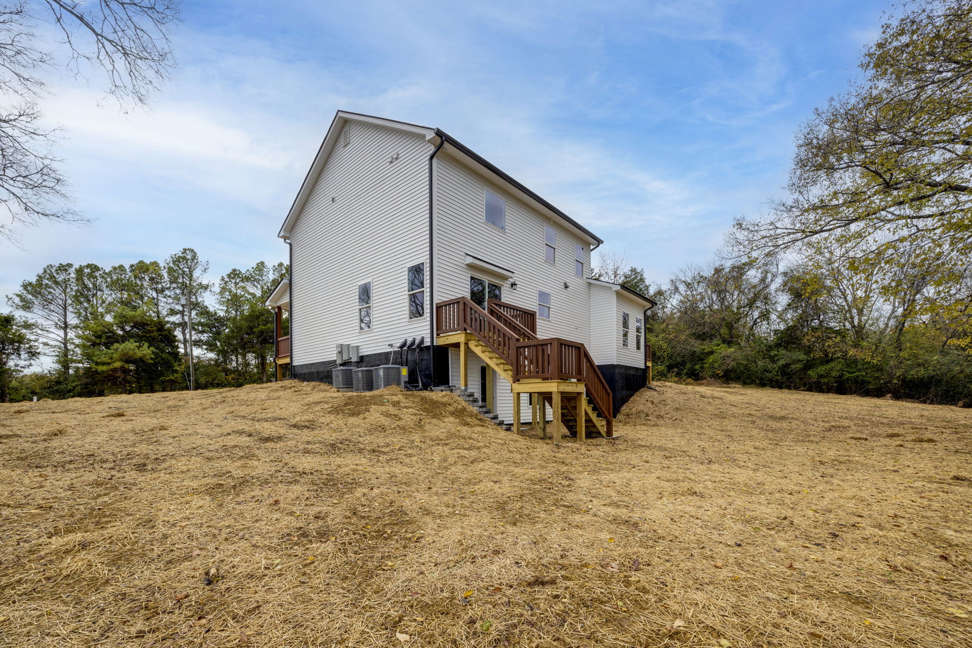 Two-story house on a grassy hill with exterior wooden staircase and deck, surrounded by trees under partly cloudy sky