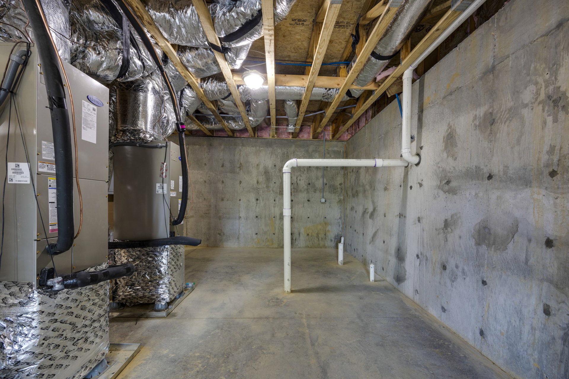 Concrete basement room with exposed pipes along ceiling and walls, plaster surfaces, and a heater unit.