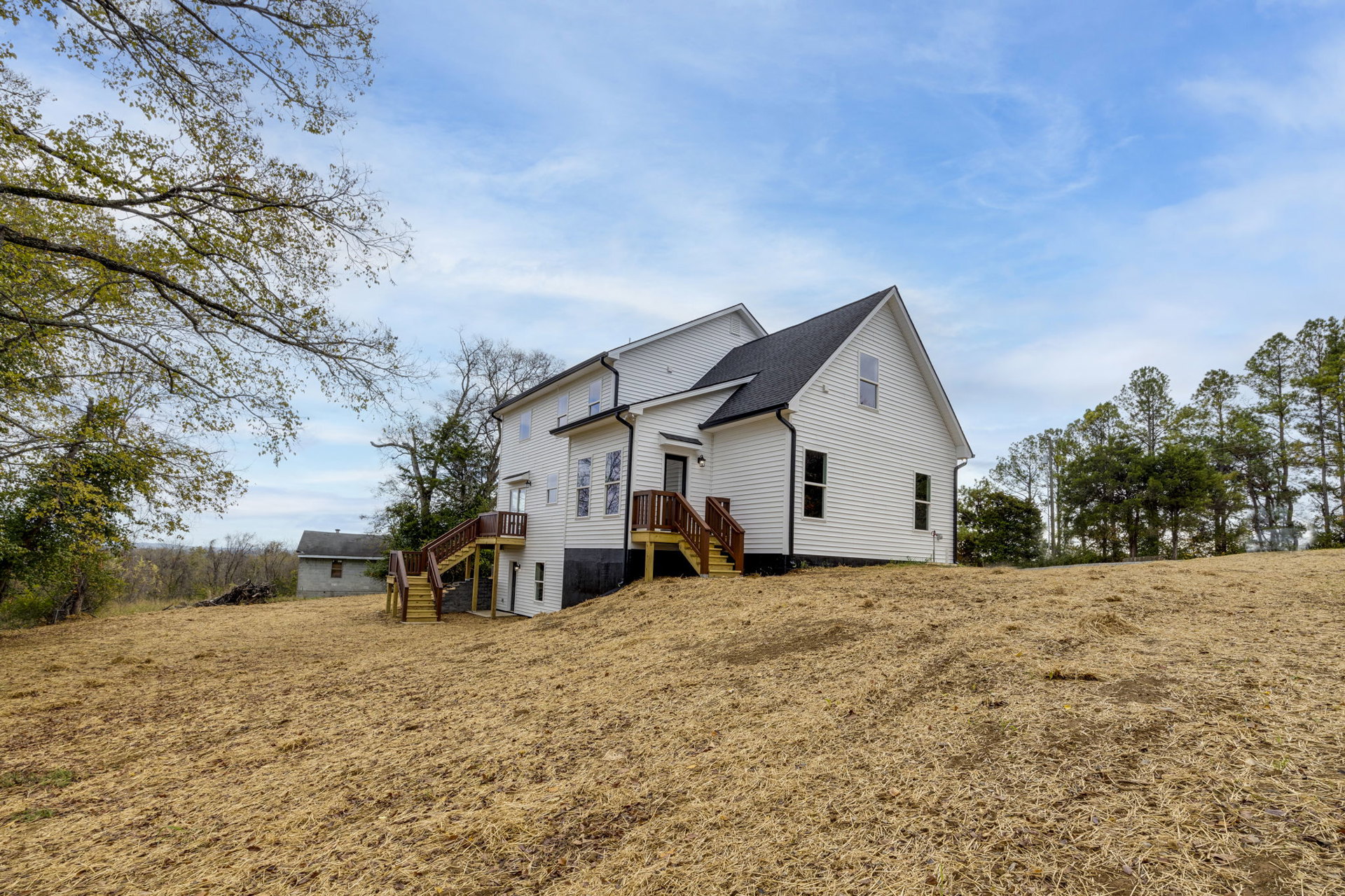 White cottage-style house on grassy hill, wooden exterior staircase, leafy tree branches, blue sky with scattered clouds