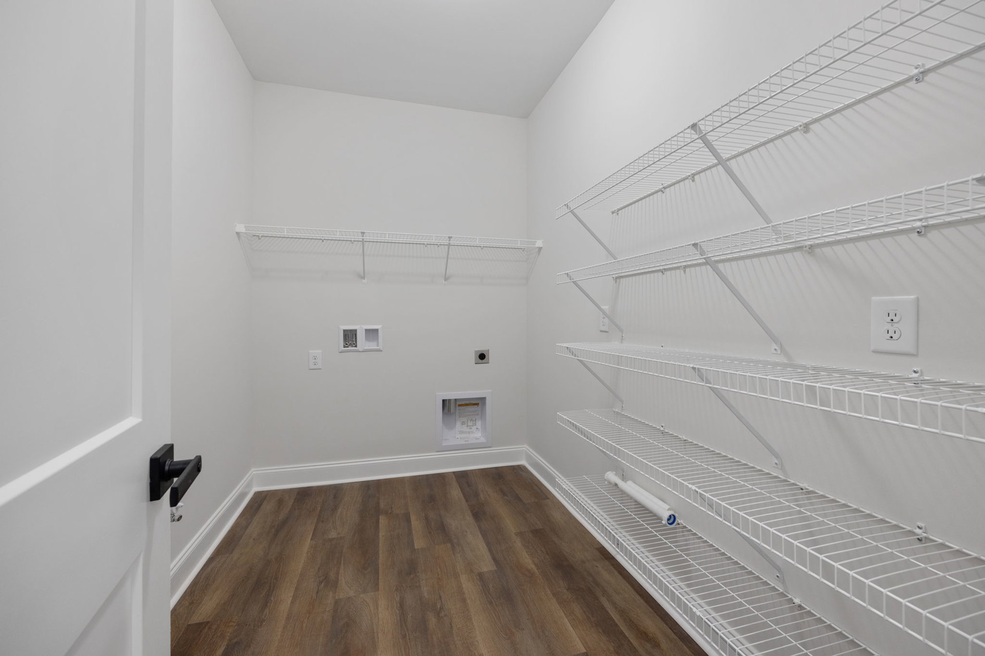 White wire shelving mounted on a plaster wall, wood flooring with white trim, close-up of a white electrical outlet, rectangular window with glass, and a white box containing
