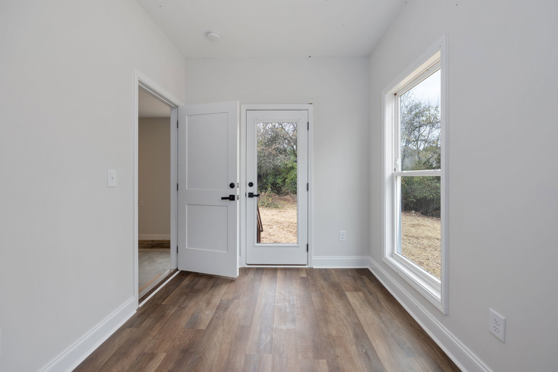 White walls, wood laminate flooring, white door with black handle, windows framing forest view, close-up of wall outlet