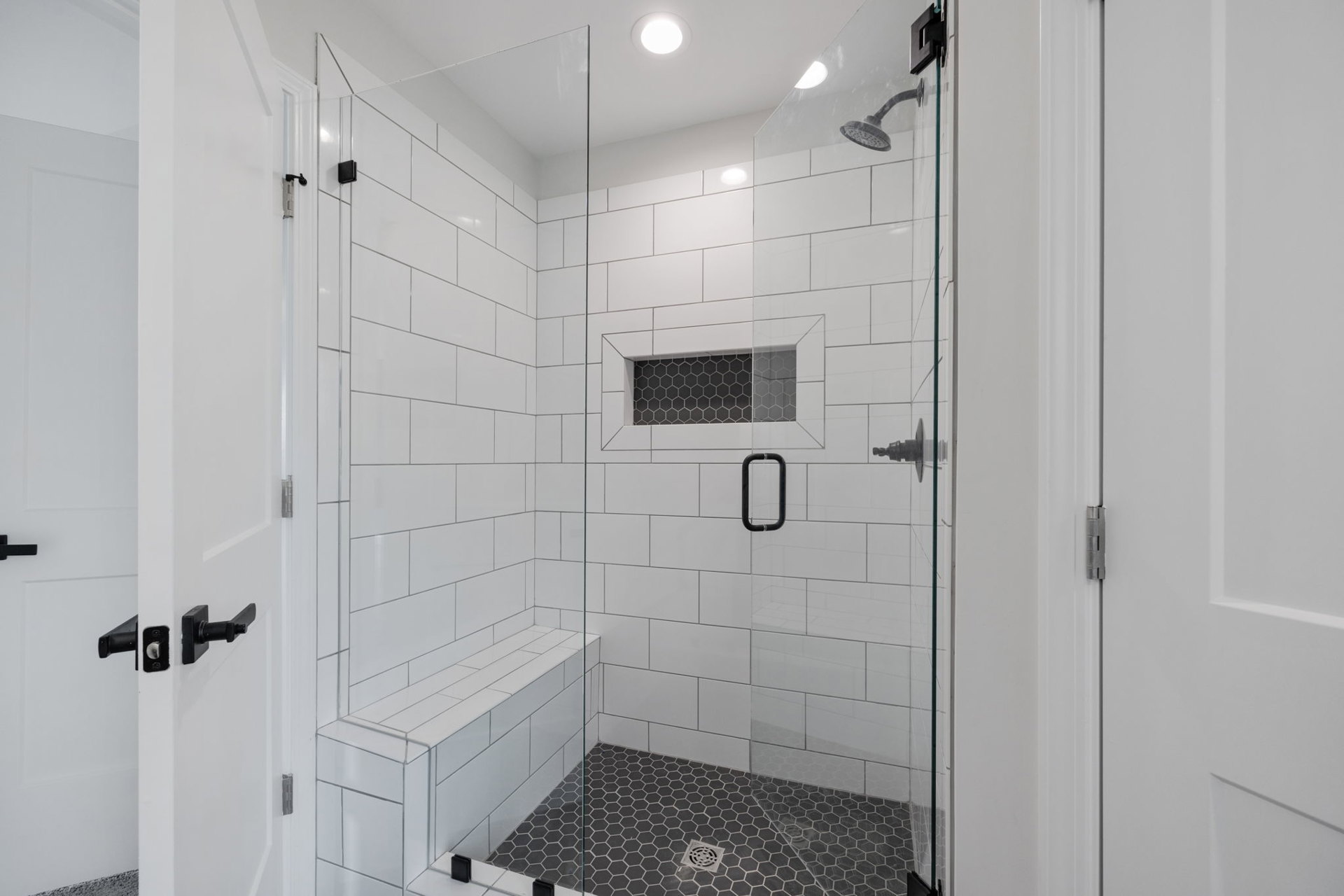 Walk-in shower with frameless glass door, white tile walls, ceiling light, black metal towel bar, floor drain, and window.
