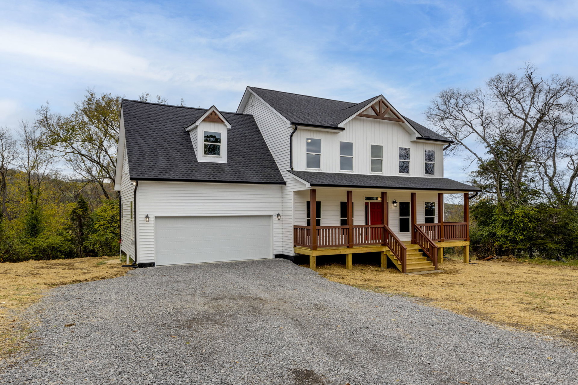 Two-story house with white garage door, red front door, wooden deck with railing, large windows illuminated from inside, leafless tree in front yard, cloudy sky overhead