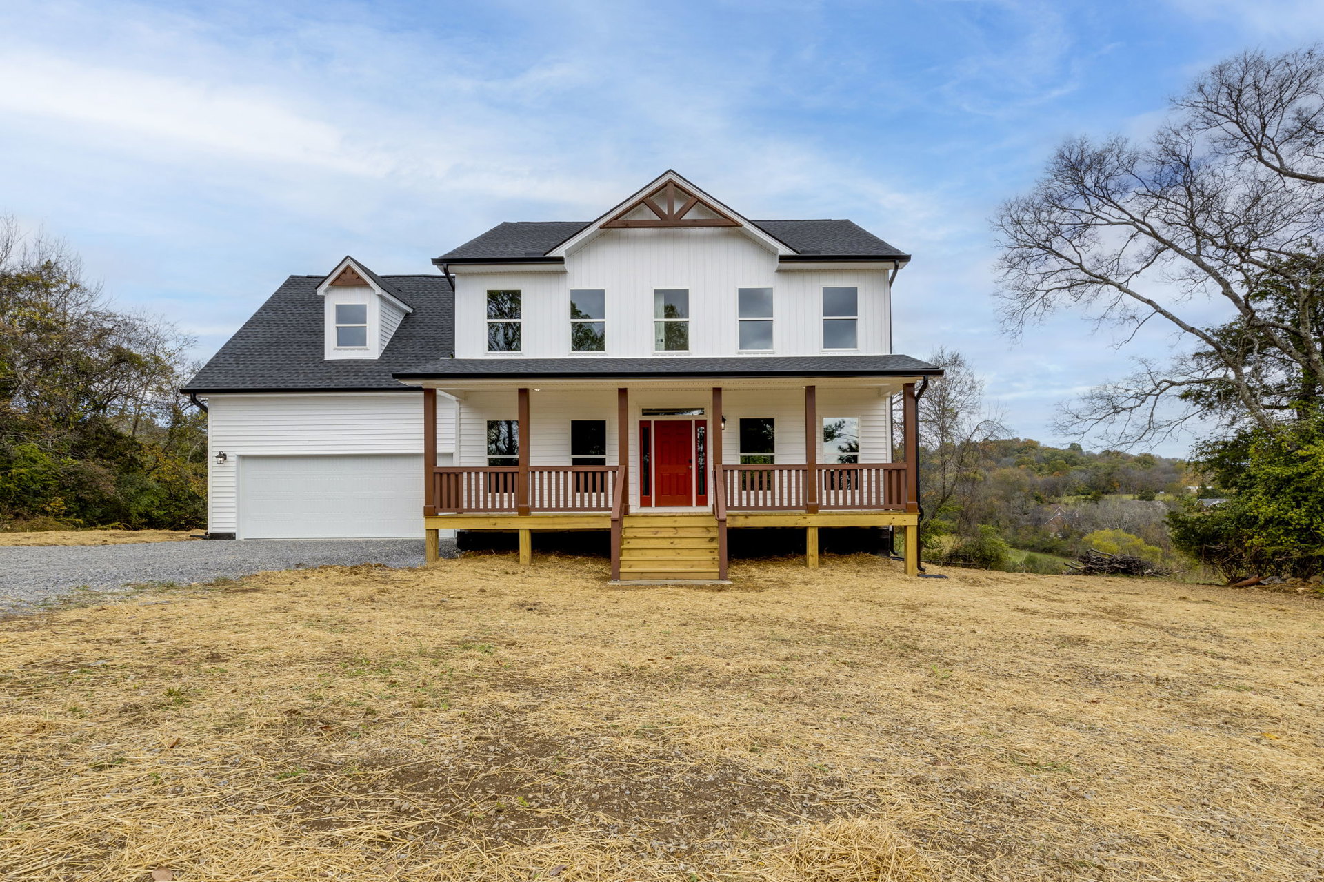 White house with red door and white trim, front porch, large grassy yard with scattered hay, white-framed windows, trees and plants under a partly cloudy sky