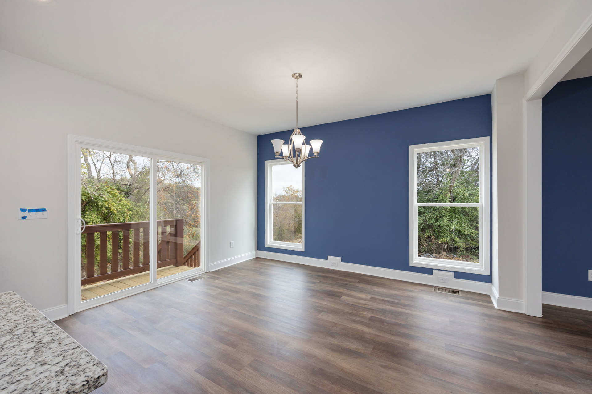 Blue-painted walls, wide plank wood flooring, marble-topped table, wooden railing, chandelier, large window with leafy trees visible outside