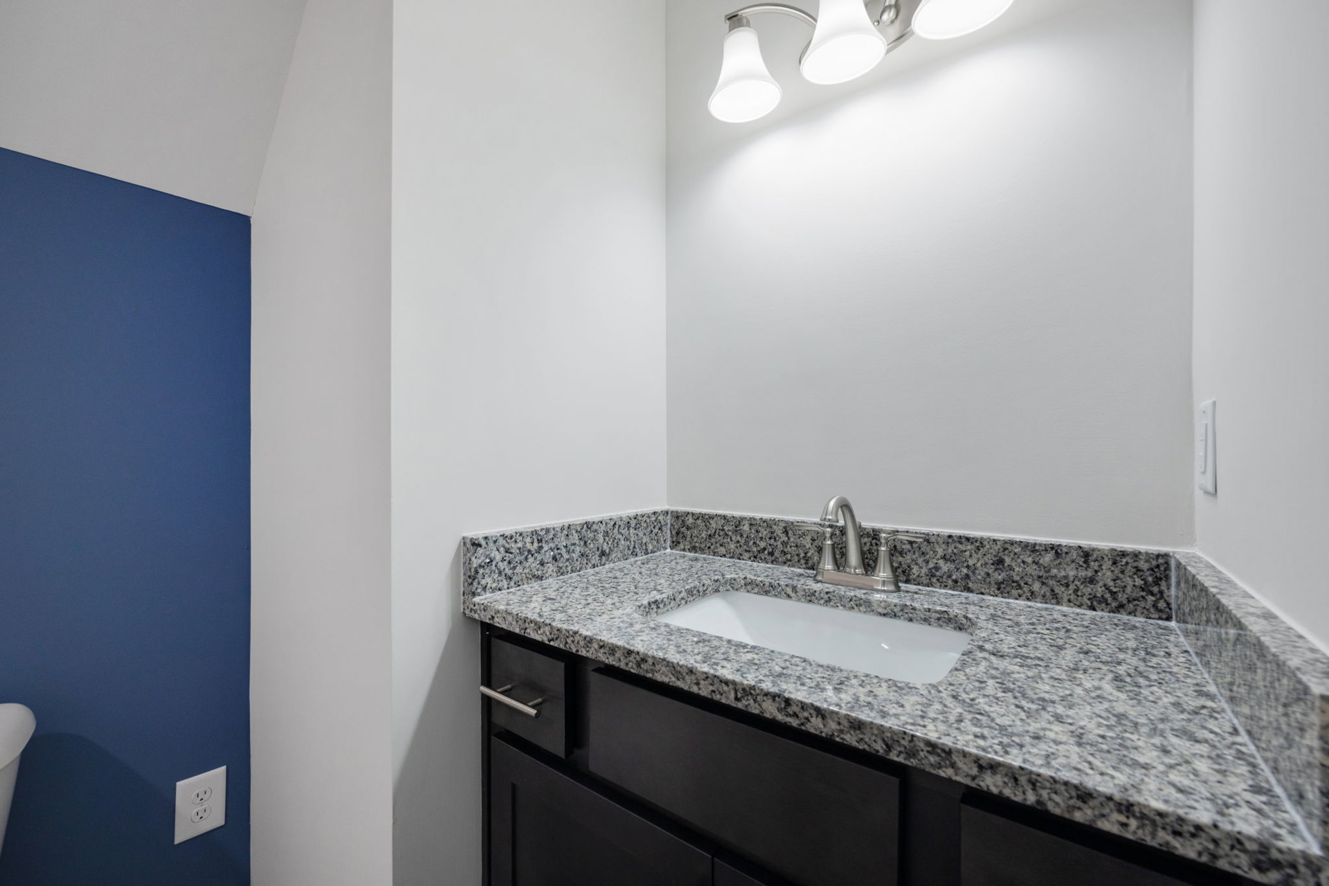 White undermount sink set in a quartz countertop, chrome faucet, light gray tile backsplash, white cabinetry, and a wall-mounted light fixture in a modern bathroom.