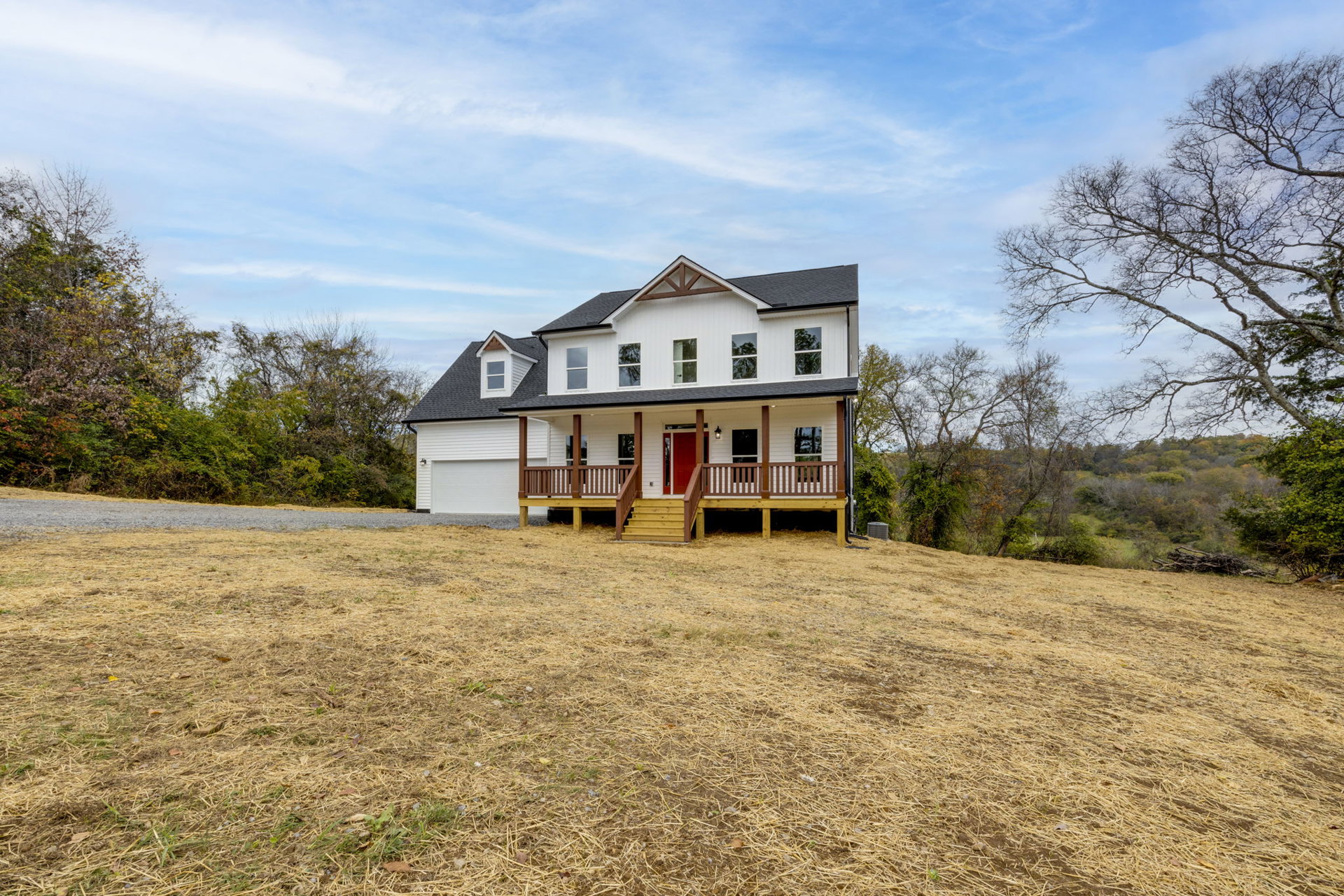 White two-story house with red front door, wide wooden deck and stairs, expansive green lawn, tree branches framing blue sky