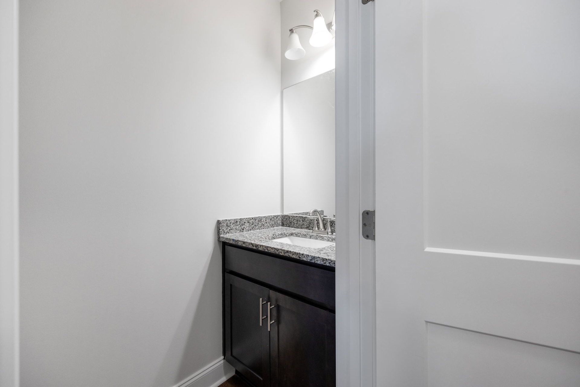 Bathroom with marble countertop, white sink, silver faucet, white cabinetry, wall-mounted mirror, two-light fixture, white door with visible hinge, and white walls.
