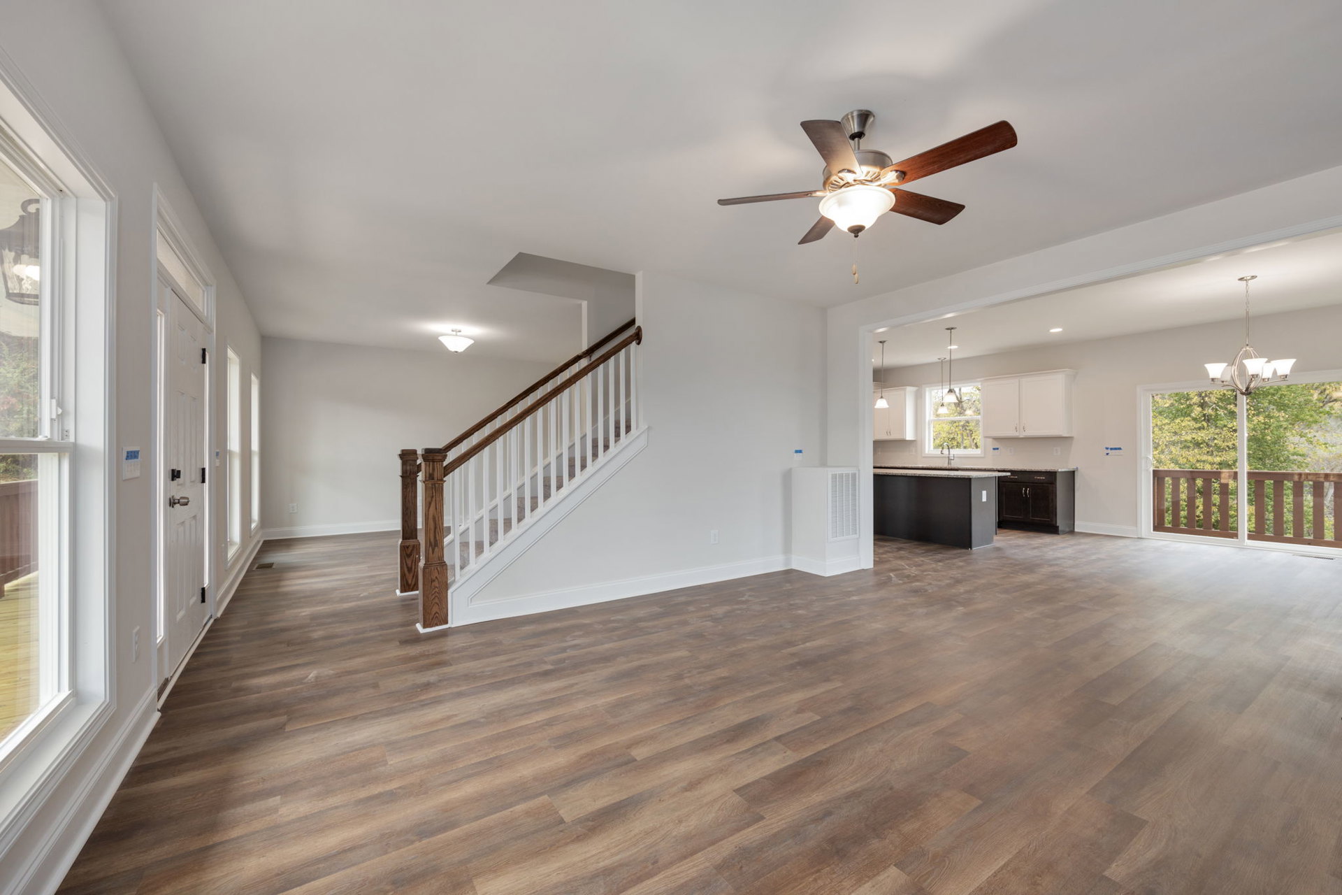 Open living area with hardwood floors, white walls, ceiling fan with light fixture, and staircase featuring wooden steps and metal railing; balcony visible through large window.