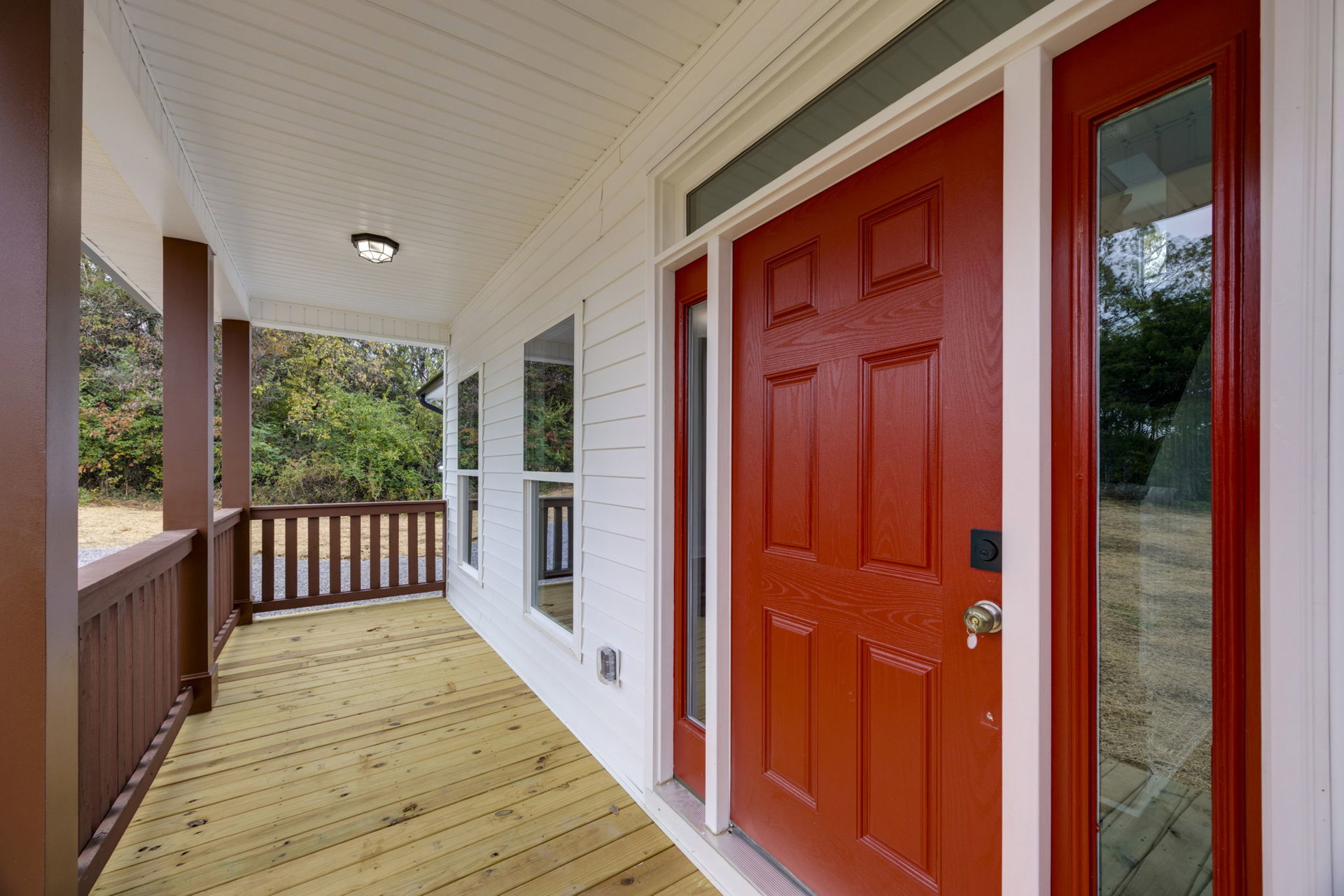 White siding house with a red front door, wooden deck and brown railing, glass window reflecting tree branches