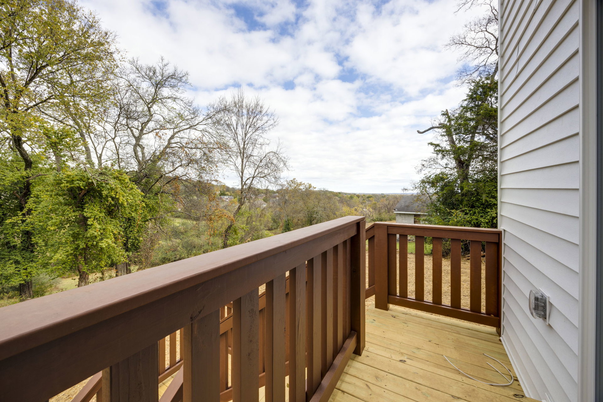 Wooden deck with brown railing overlooking leafy trees and blue sky with scattered clouds