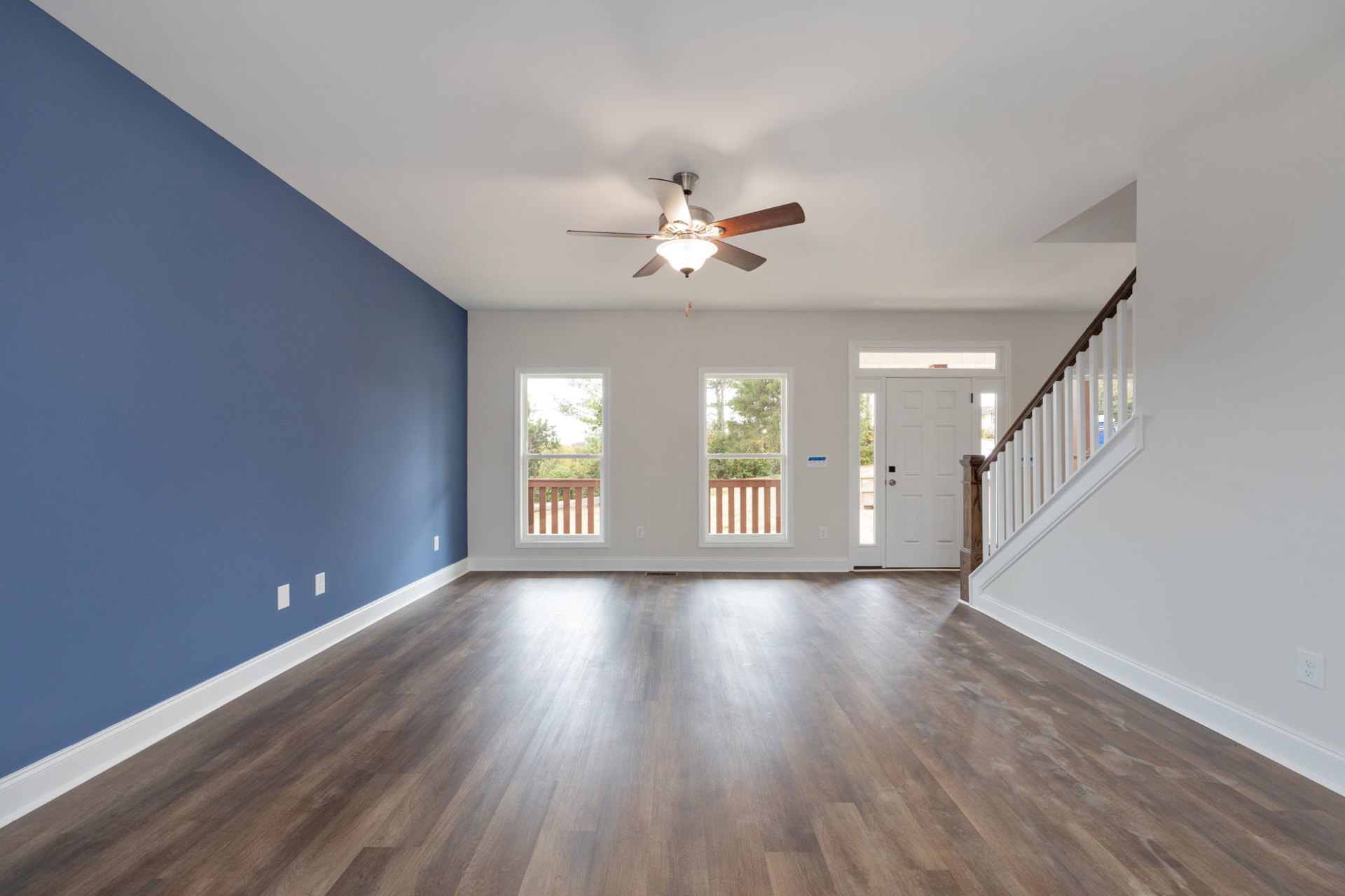 Hardwood floored room with blue walls, ceiling fan with light, wooden staircase, and large window overlooking trees