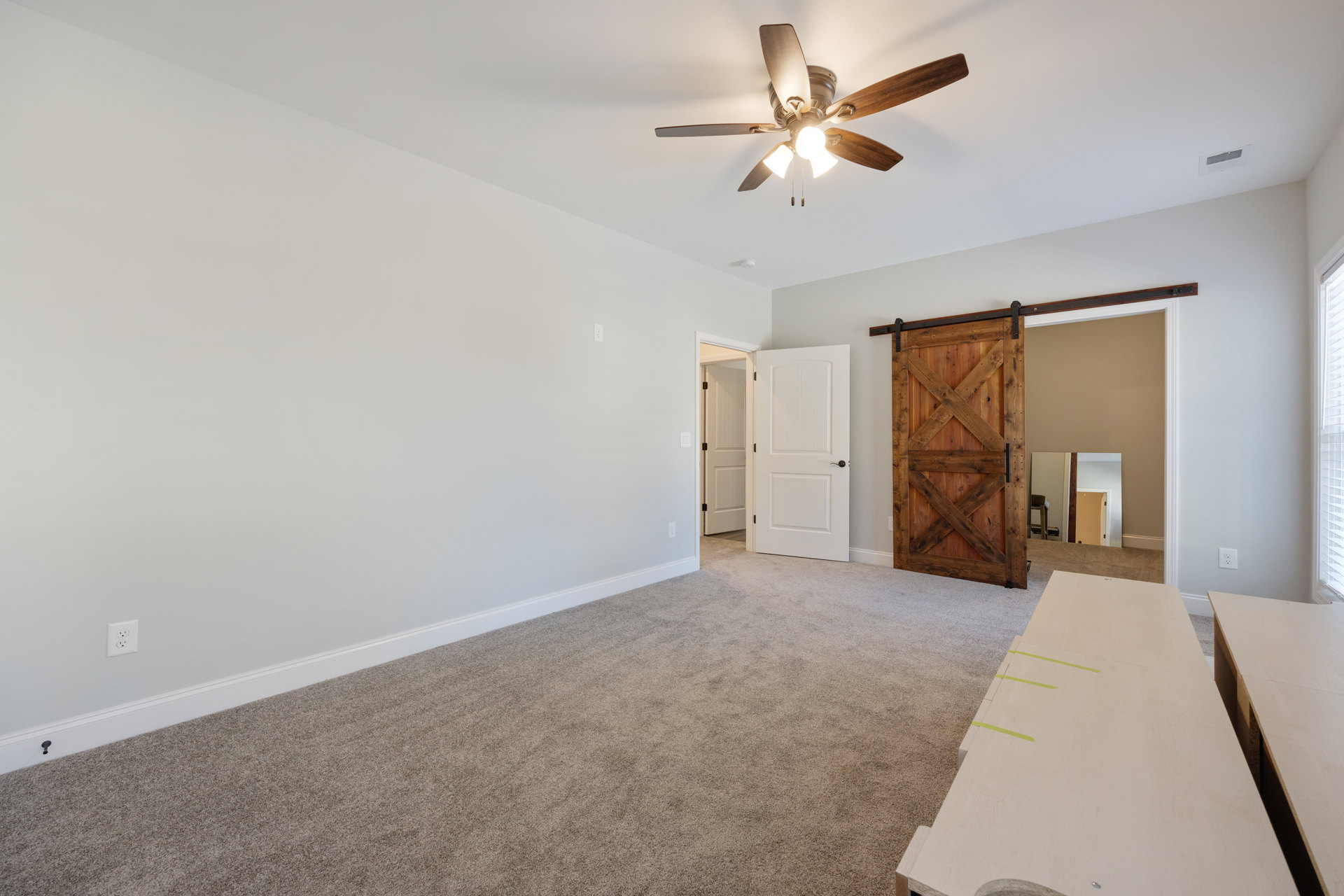Ceiling fan with light fixture, wooden door featuring a cross, white door with black handle, table marked with green tape, plaster walls, and light flooring in a residential
