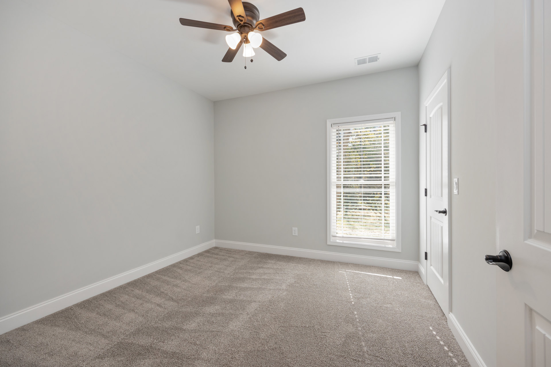 Neutral-toned carpeted room with white walls, ceiling fan with light fixture, window fitted with horizontal blinds, and door featuring a brushed metal handle