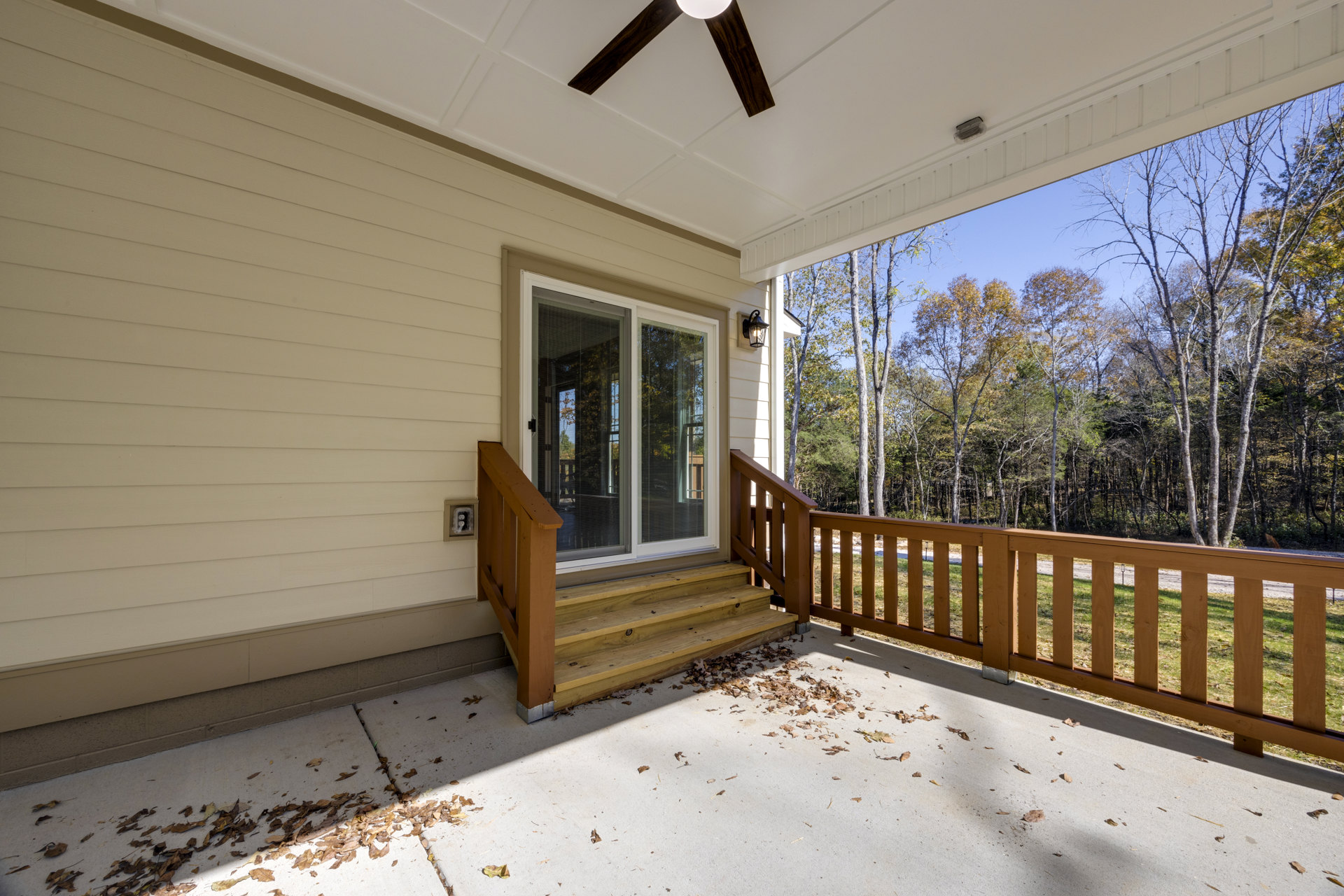 Covered porch with wooden deck, ceiling fan, sliding glass door with blinds, window with blinds, and leaves scattered near the railing