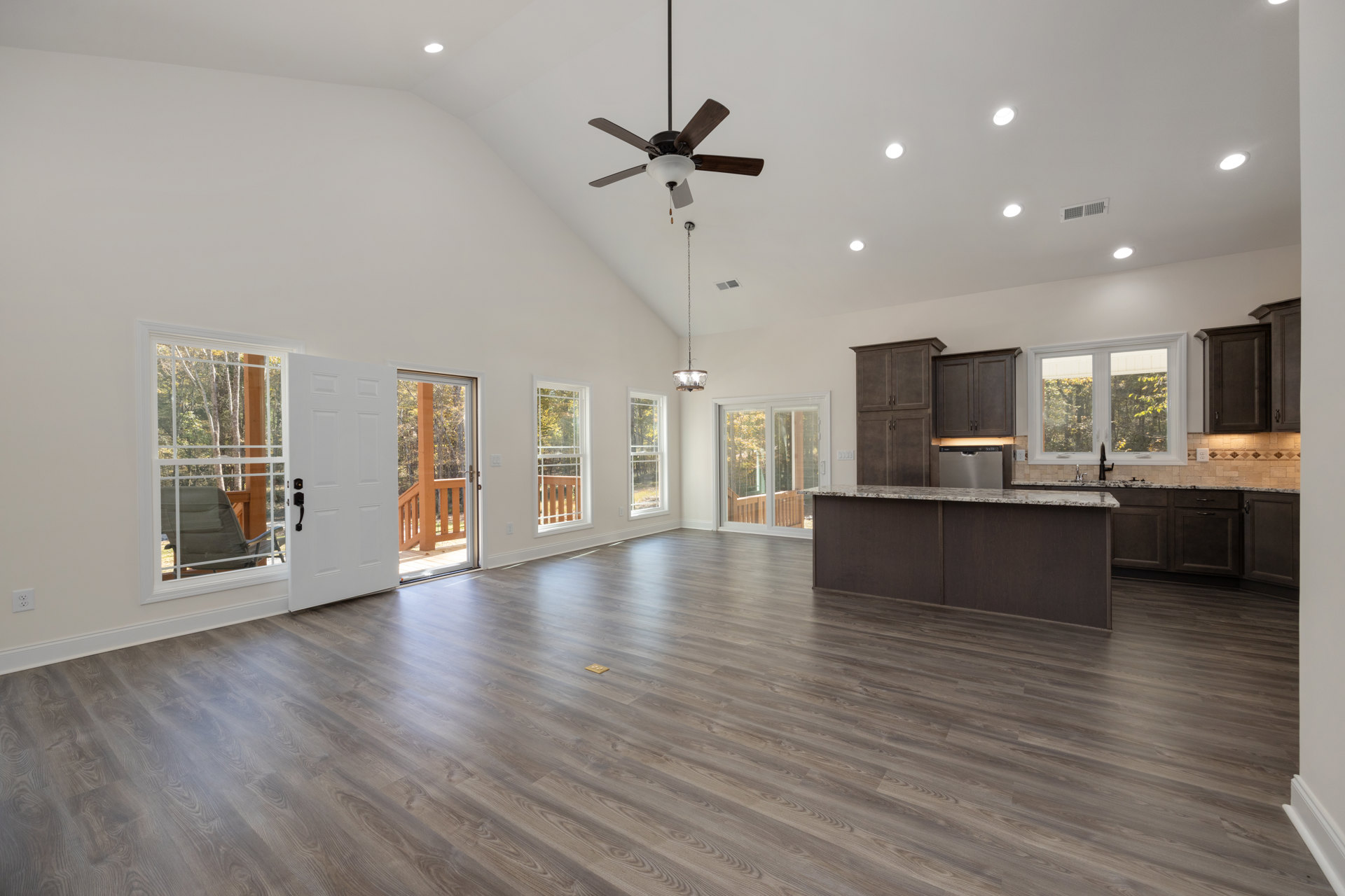 Open-concept kitchen with wood flooring, white-framed windows overlooking trees, ceiling fan with light fixture, and neutral walls.