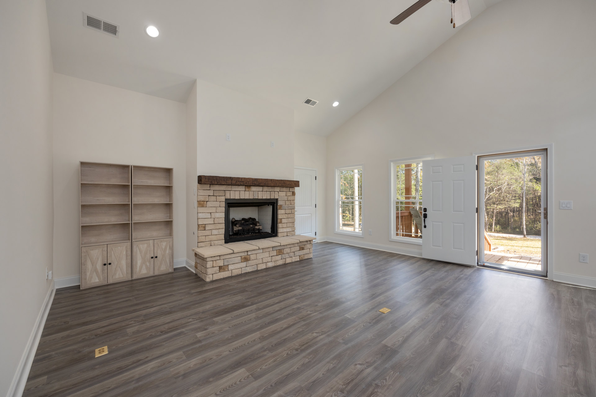 Living room with wood flooring, plaster walls, built-in wooden shelves with doors, large window overlooking trees and grass, and a fireplace burning wood logs.