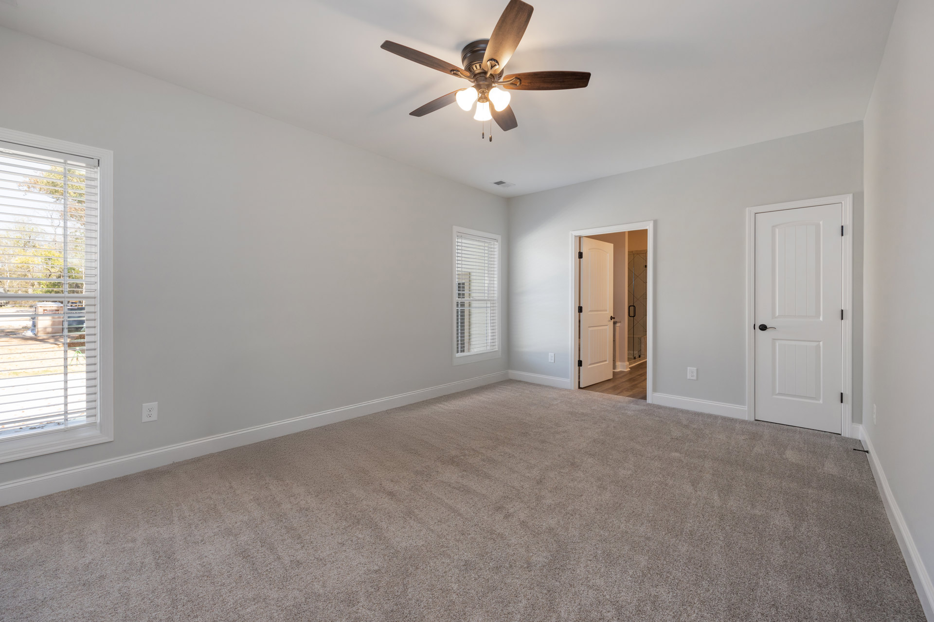 Carpeted bedroom with ceiling fan and light, white door with black handle, glass shower door, window with white blinds, trees visible outside