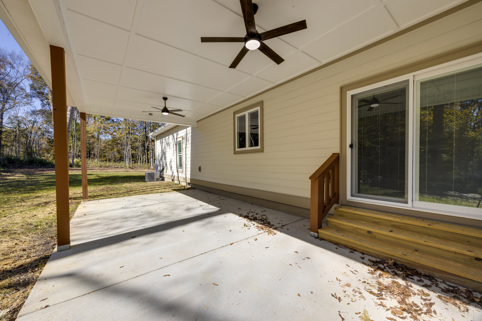 Front porch with wooden railing, ceiling fan with light fixture, white-framed windows featuring black handles and side blinds, surrounded by trees and landscaped ground.