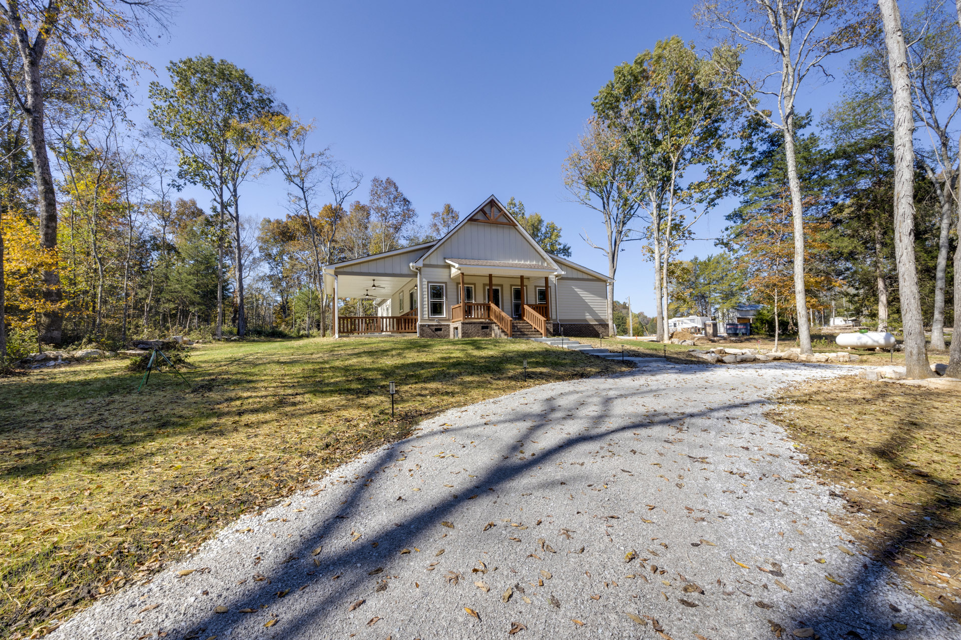 Two-story house with white siding, covered front porch, wooden railing, concrete driveway, mature trees, autumn leaves scattered on grass, clear blue sky