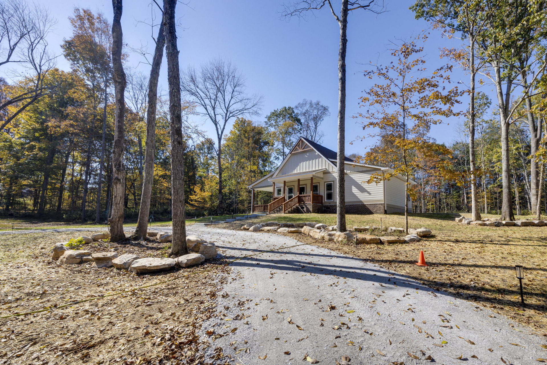 Two-story house with brown wooden porch and staircase, surrounded by deciduous trees with yellow and green autumn leaves, gravel path bordered by rocks and fallen leaves in front
