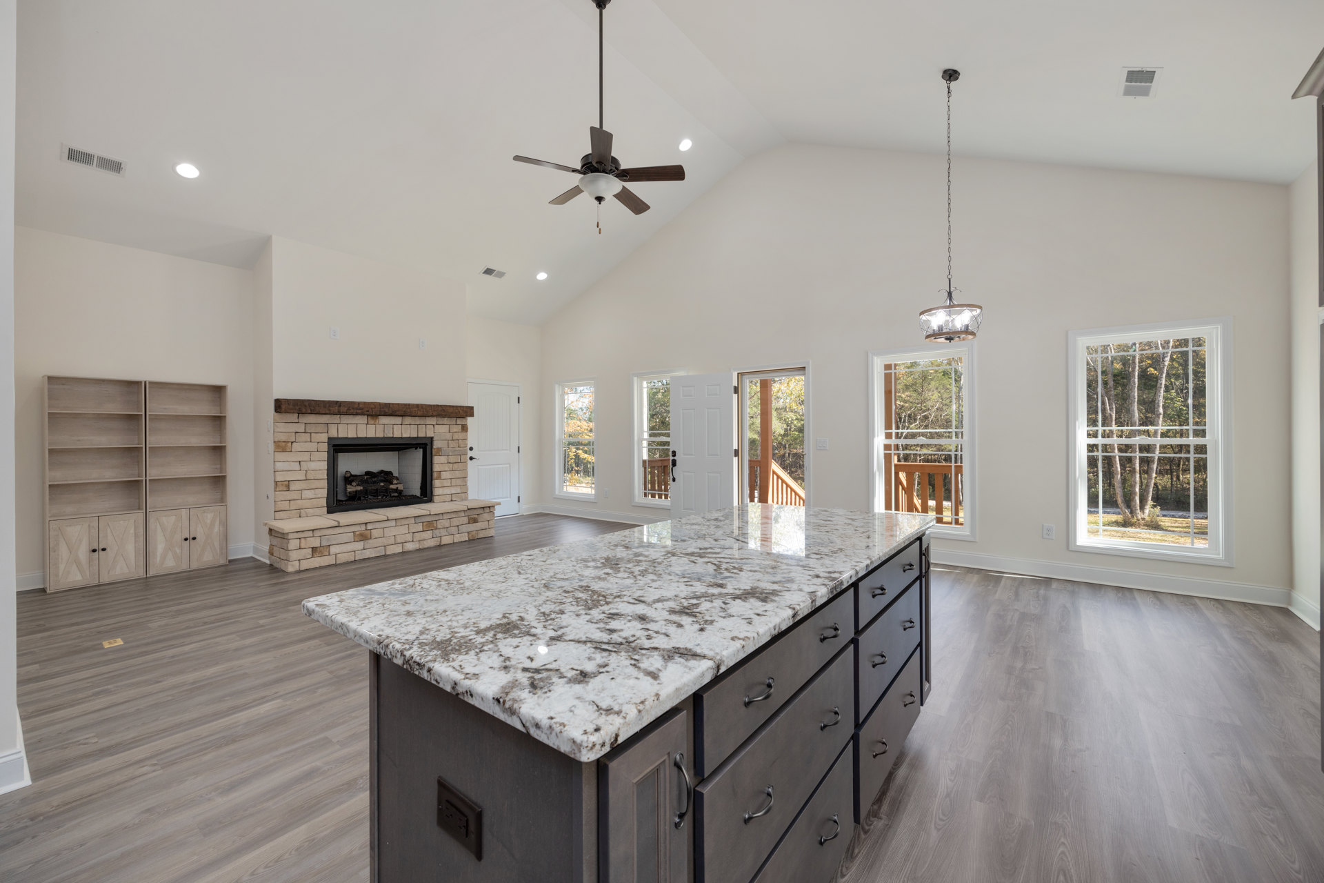 Marble-topped kitchen island in spacious room, fireplace with burning log, window showing trees, two wall shelves, ceiling fan with light fixture, wood cabinetry and flooring