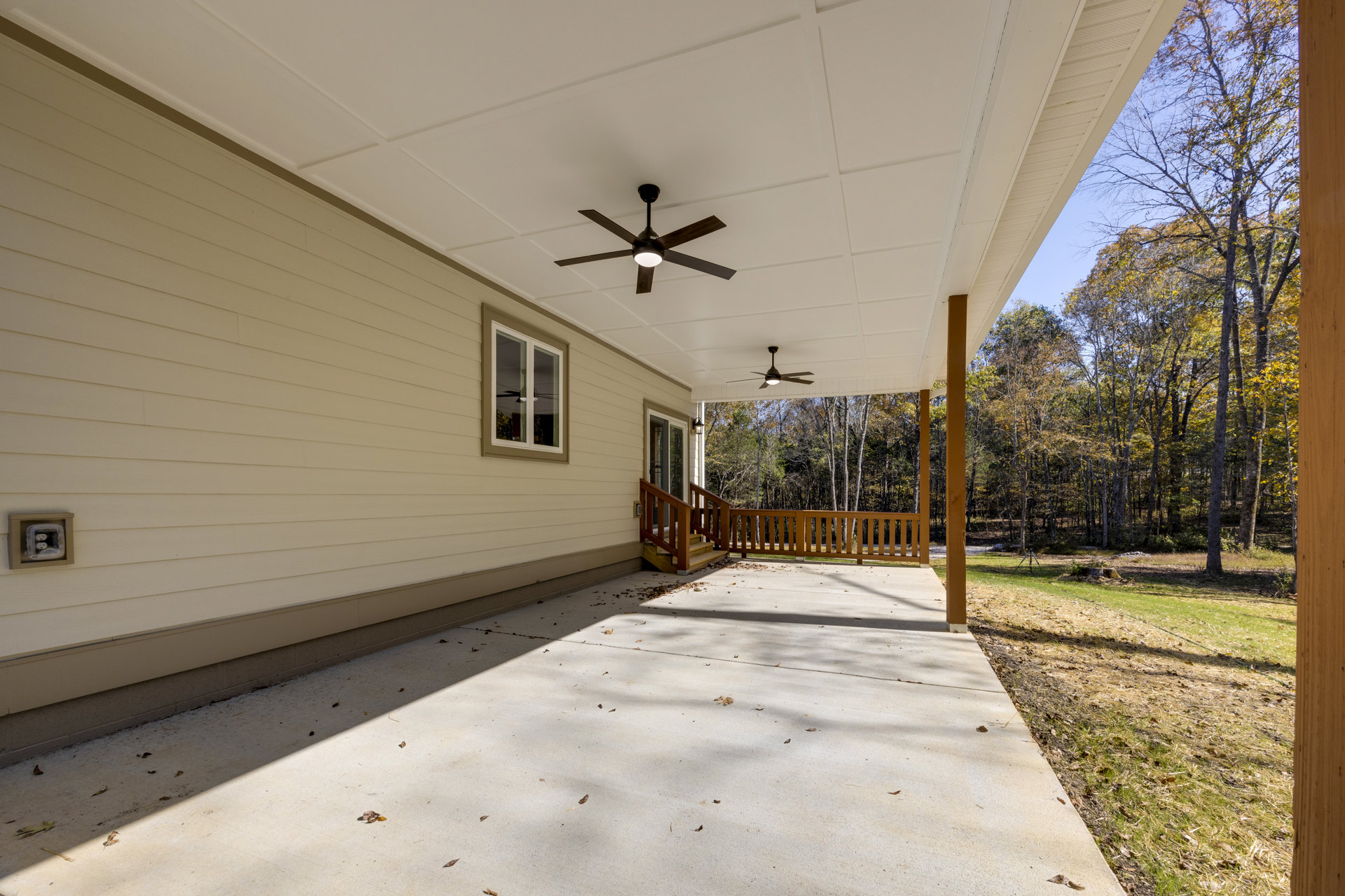 Wood deck with wooden railing, ceiling fan with light fixture above, large windows, concrete patio below, leafy trees in background