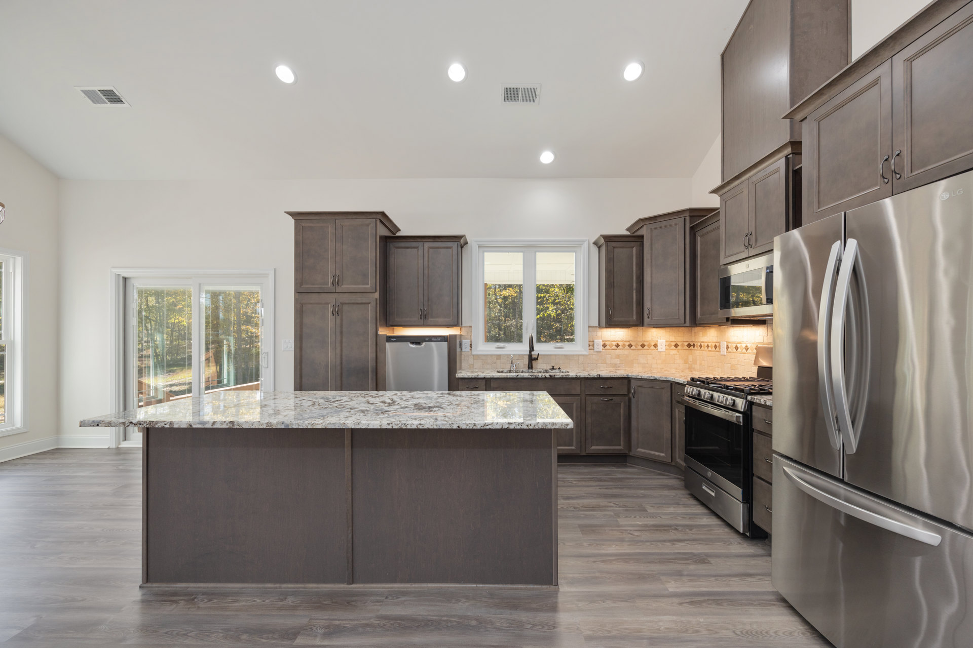 Marble island with waterfall edge, stainless steel refrigerator and stove, wood base cabinetry, white tile backsplash, large window overlooking trees, light hardwood flooring