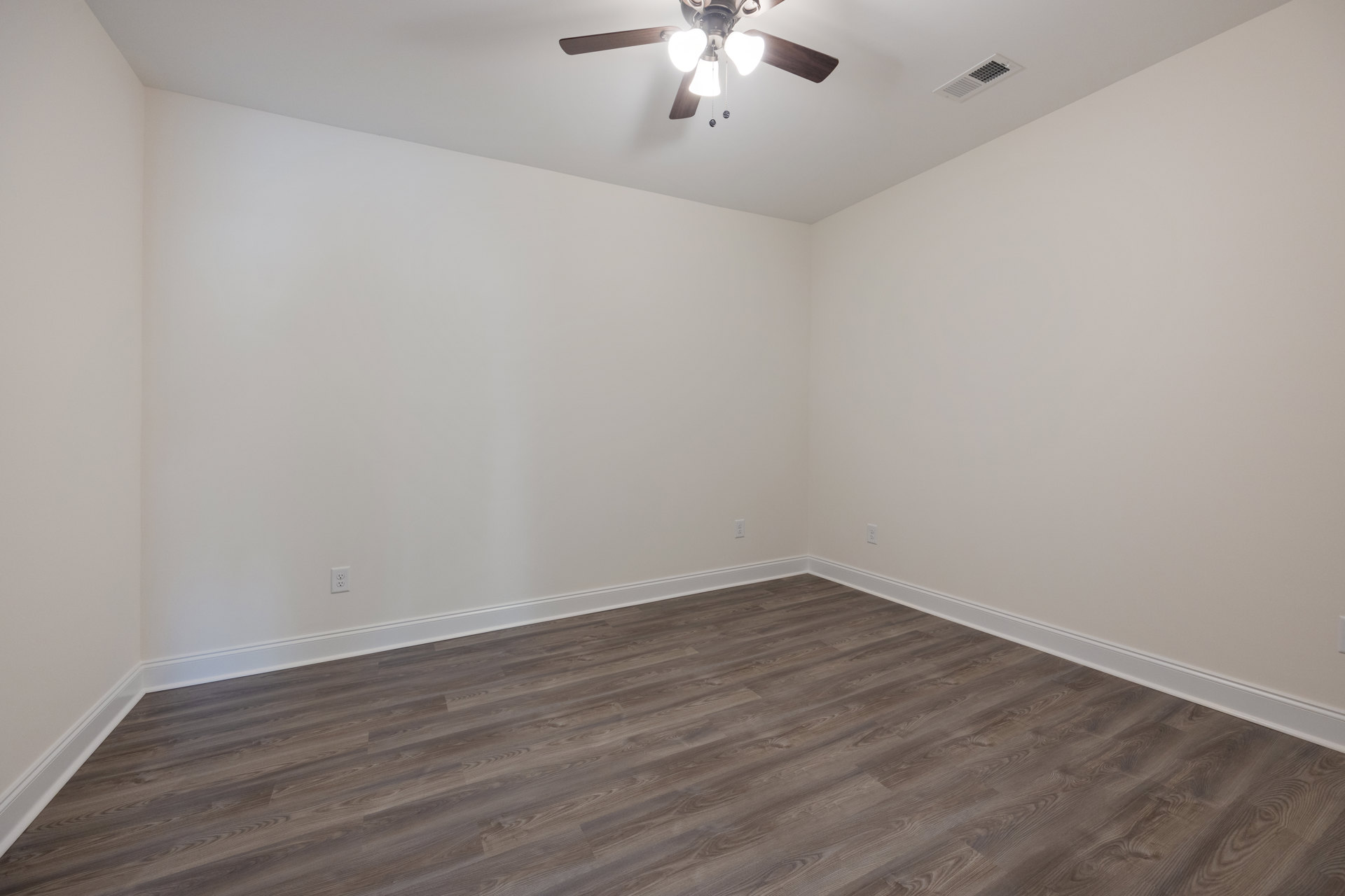 Ceiling fan with lights mounted on white plaster ceiling, wood flooring with visible grain pattern, wall molding, and ceiling vent in a residential room