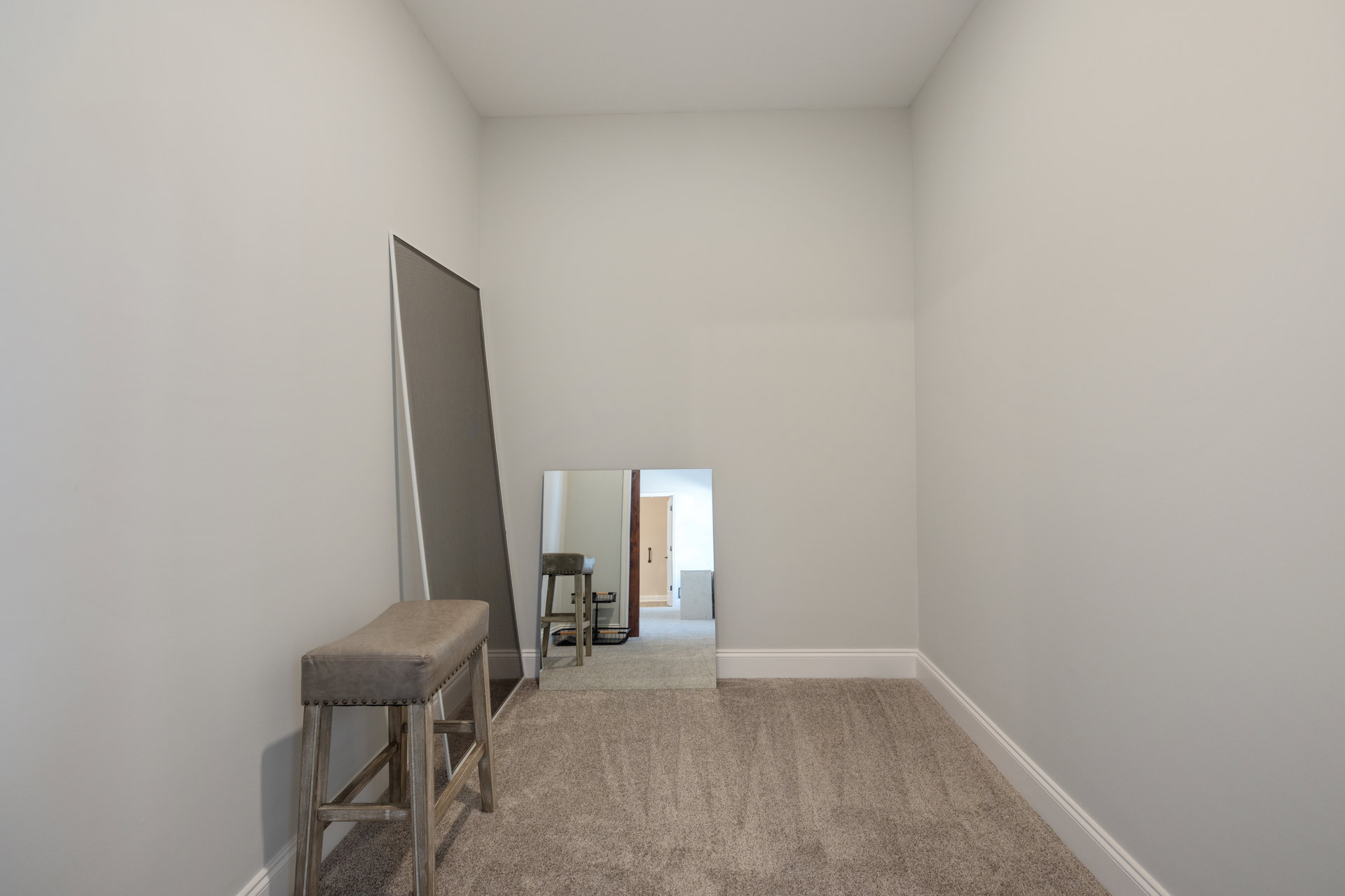 Large rectangular mirror mounted on a white wall, black background reflection, white door partially visible, light-colored carpet with square pattern, small stool placed near