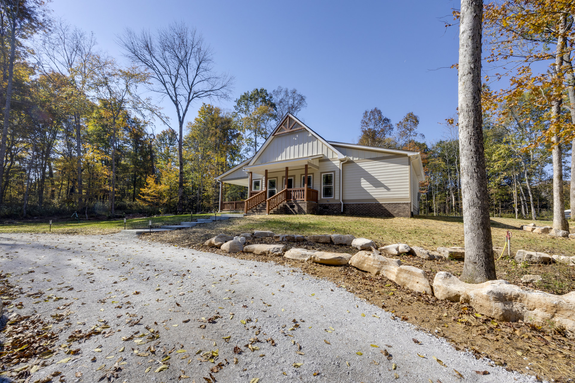 Two-story house with covered porch, white siding, and front stairs, surrounded by autumn trees and a gravel driveway edged with rocks